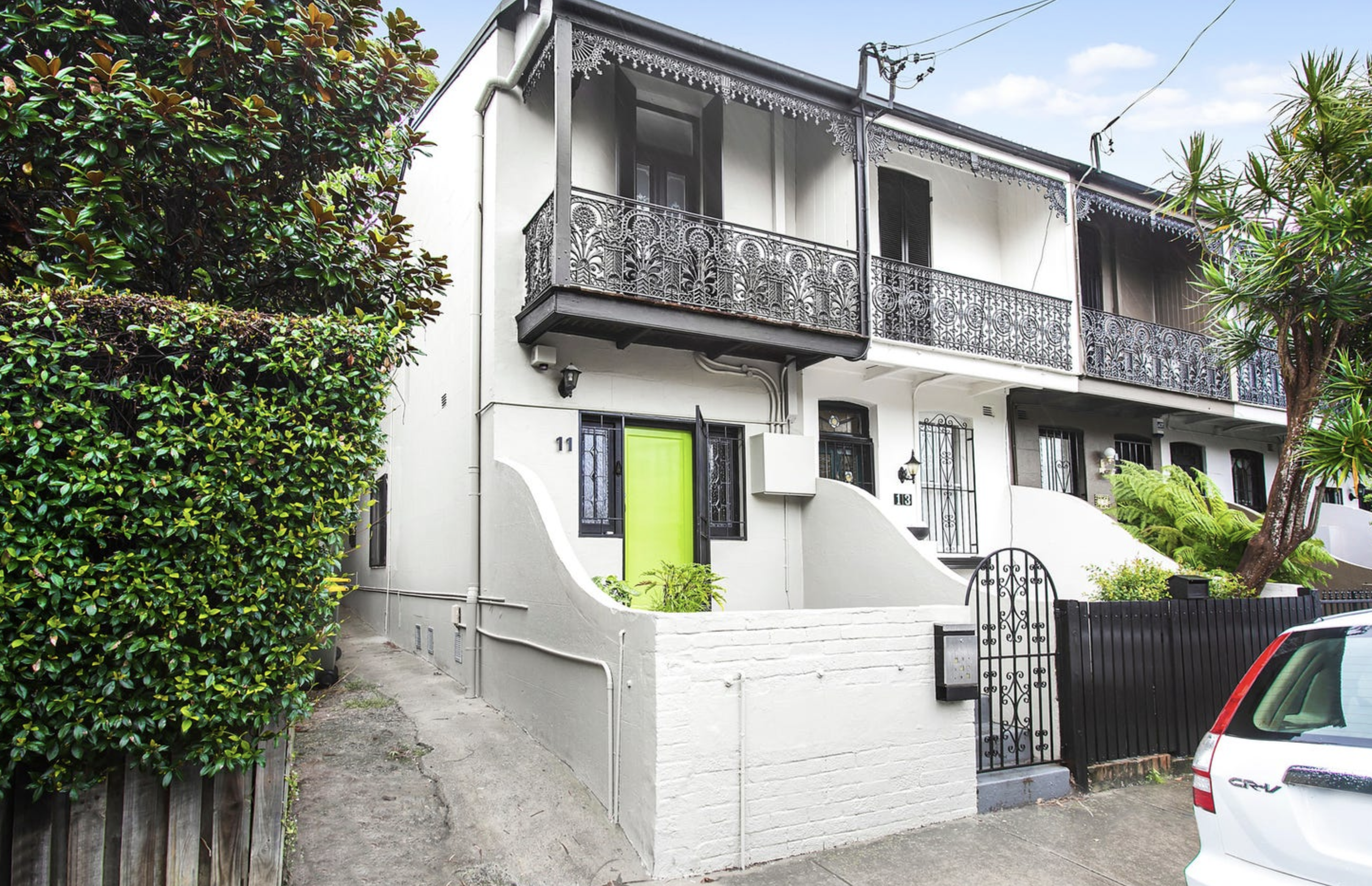 A row of attached white residential buildings with black wrought-iron balconies and black gates, greenery, and a sidewalk in front.