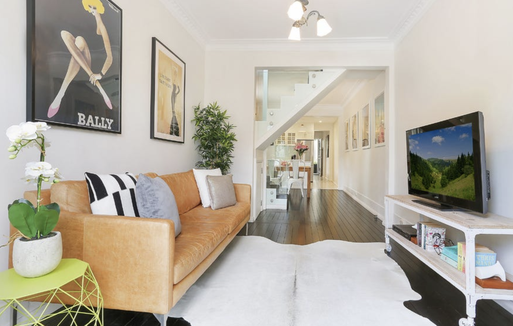 Living room with orange sofa, wall art, potted plant, TV on white stand, and dining area in background.