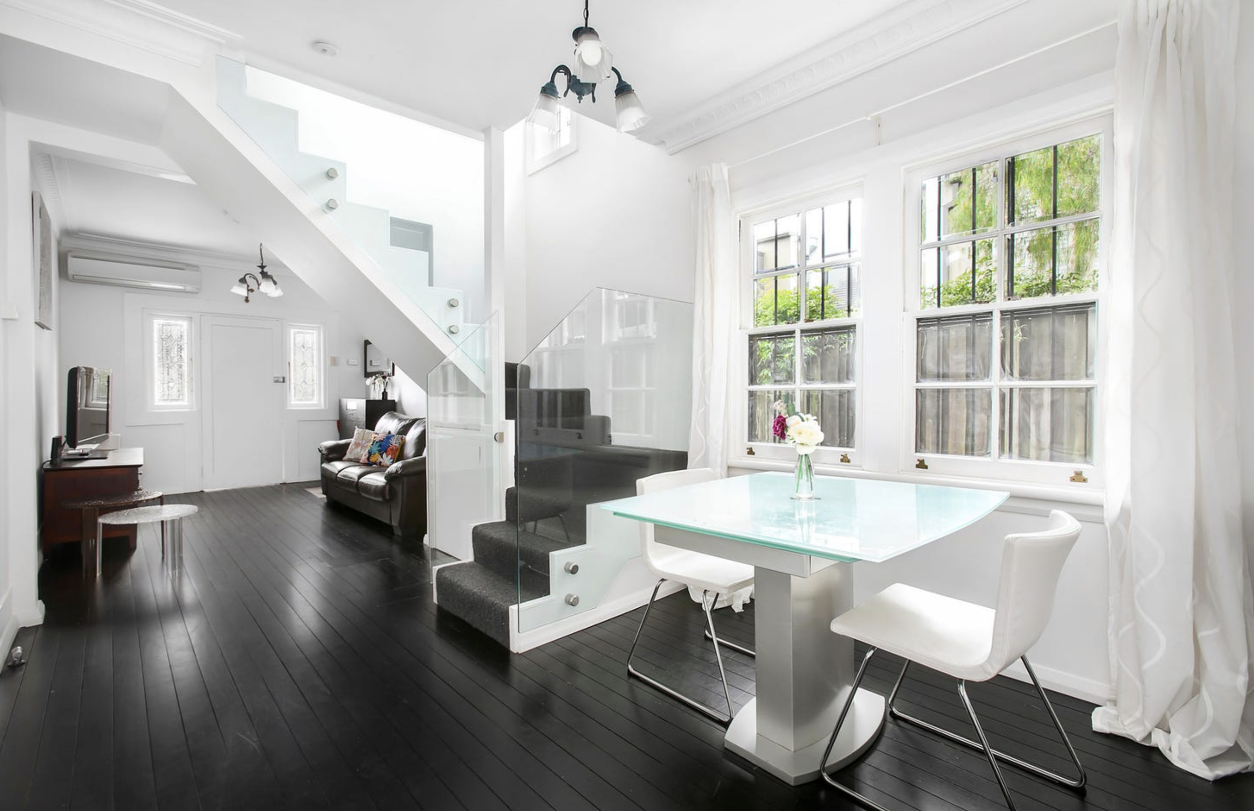 Bright living room with large windows, white walls, black flooring, modern glass dining table, white chairs, sofa, staircase with glass railing, and a chandelier.