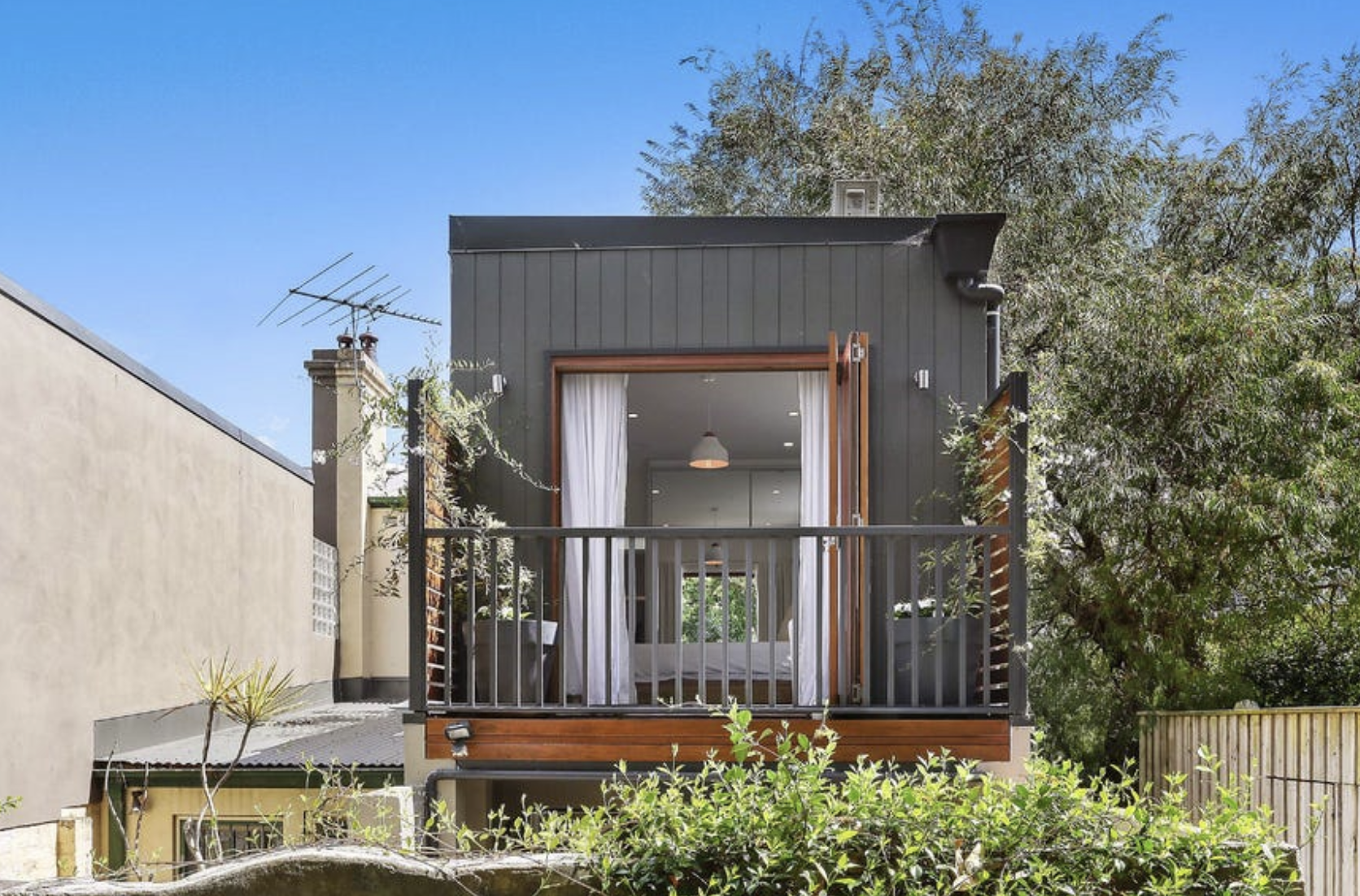 A modern two-story house with a small balcony, black exterior walls, and a sliding door with white curtains, surrounded by greenery and a clear blue sky.