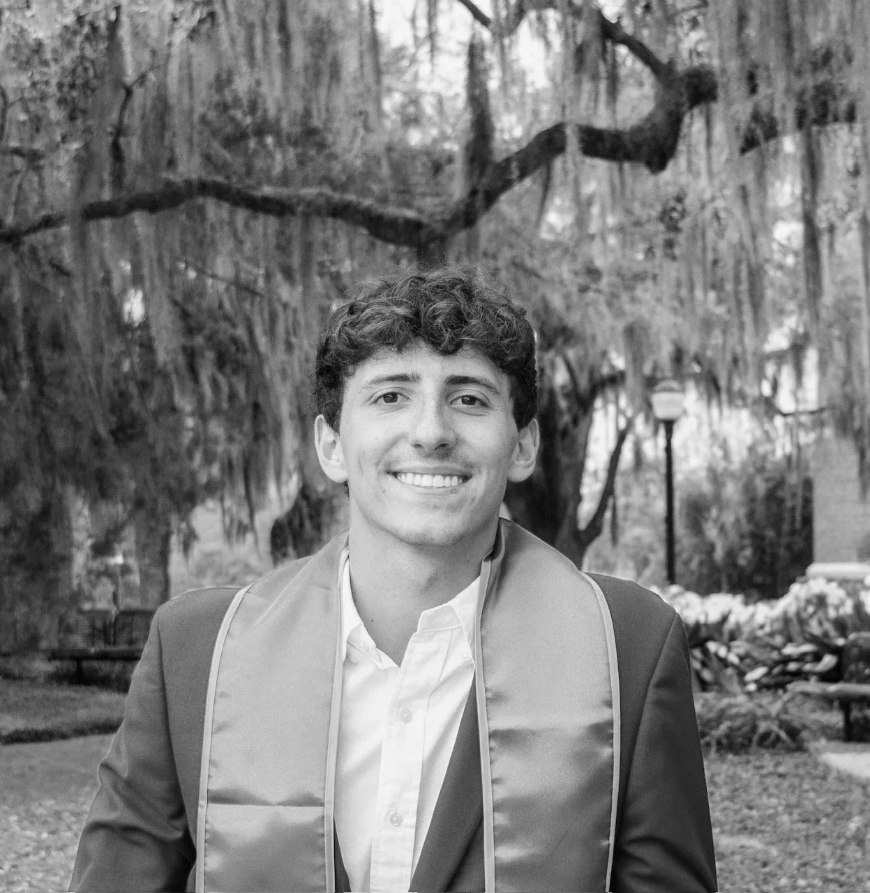 A young man with curly hair smiling in a suit and tie outdoors with trees and a lamp post in the background.