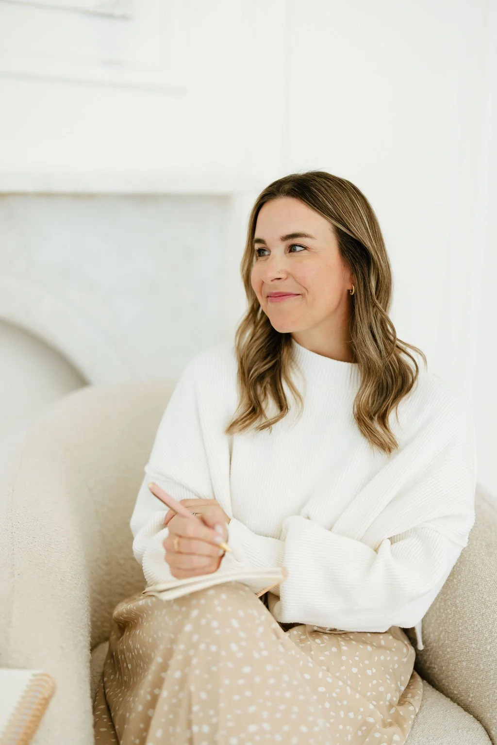 Kelly sits in a chair with a journal and pen in hand, looking off into the distance.