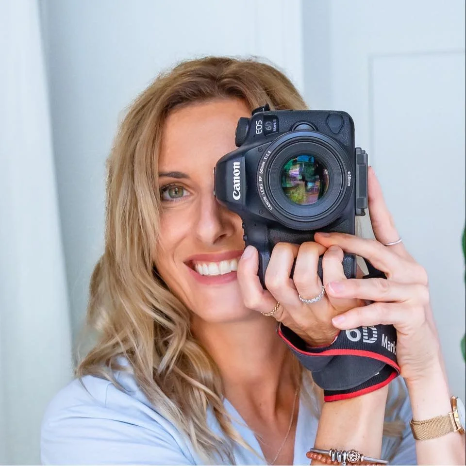 Mujer con cabello rubio y sonrisa, tomando una selfie con una cámara réflex Canon en un interior.