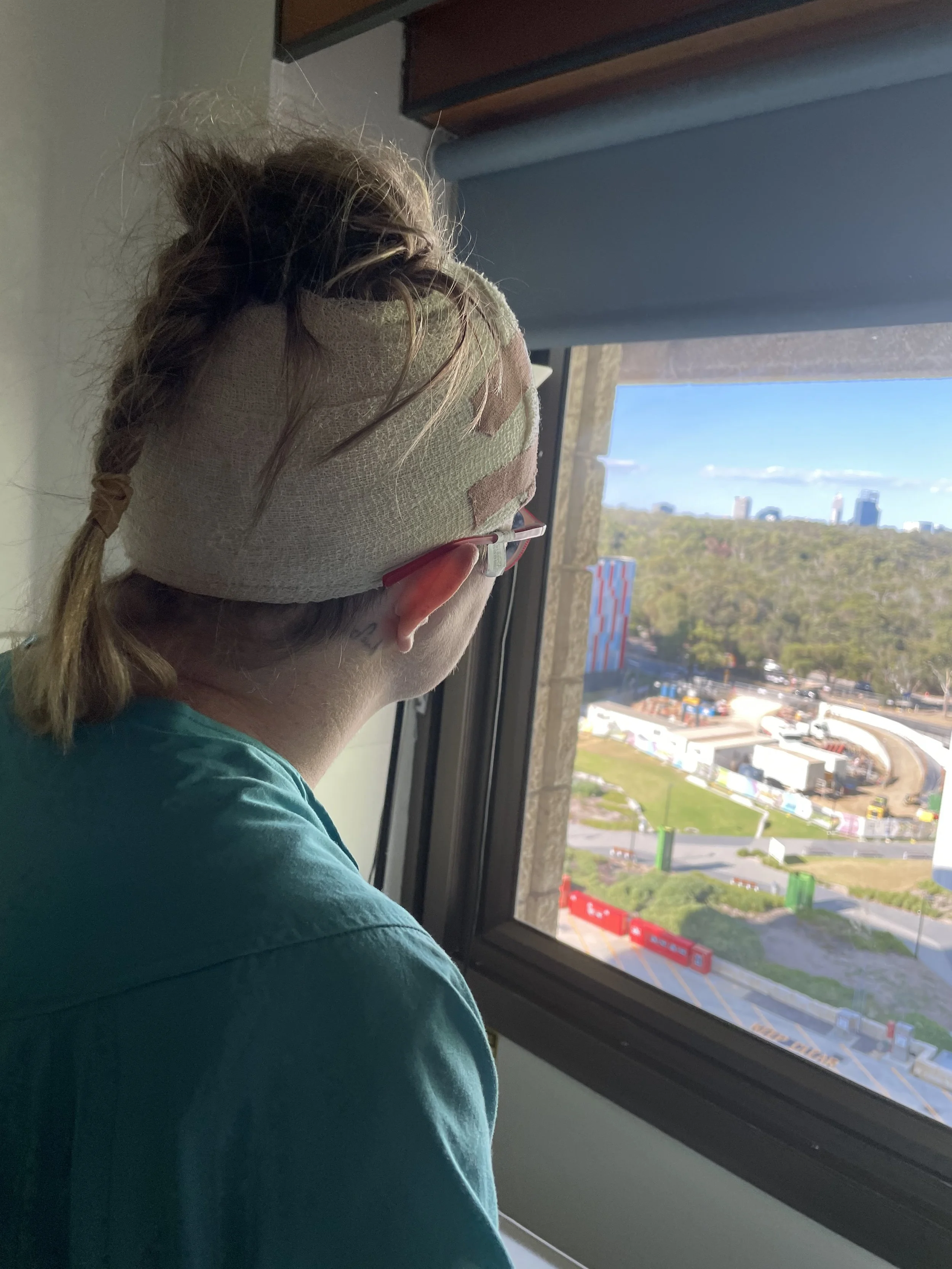 Hannah staring out her hospital window. She is wearing a greenish gown and her head is heavily bandaged from brain surgery. She is now deaf.