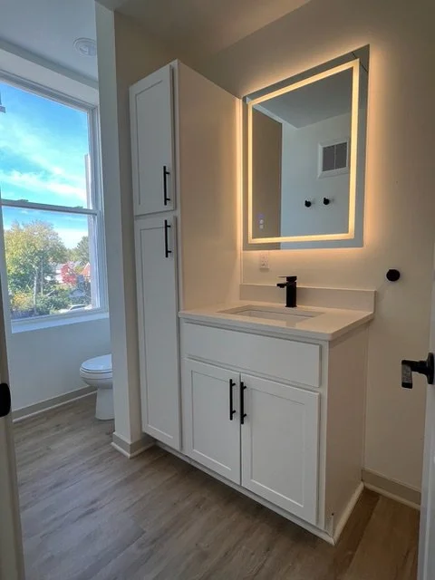 Modern bathroom vanity with a mirror, black fixtures, white cabinets, and a window showing a view of trees outside.