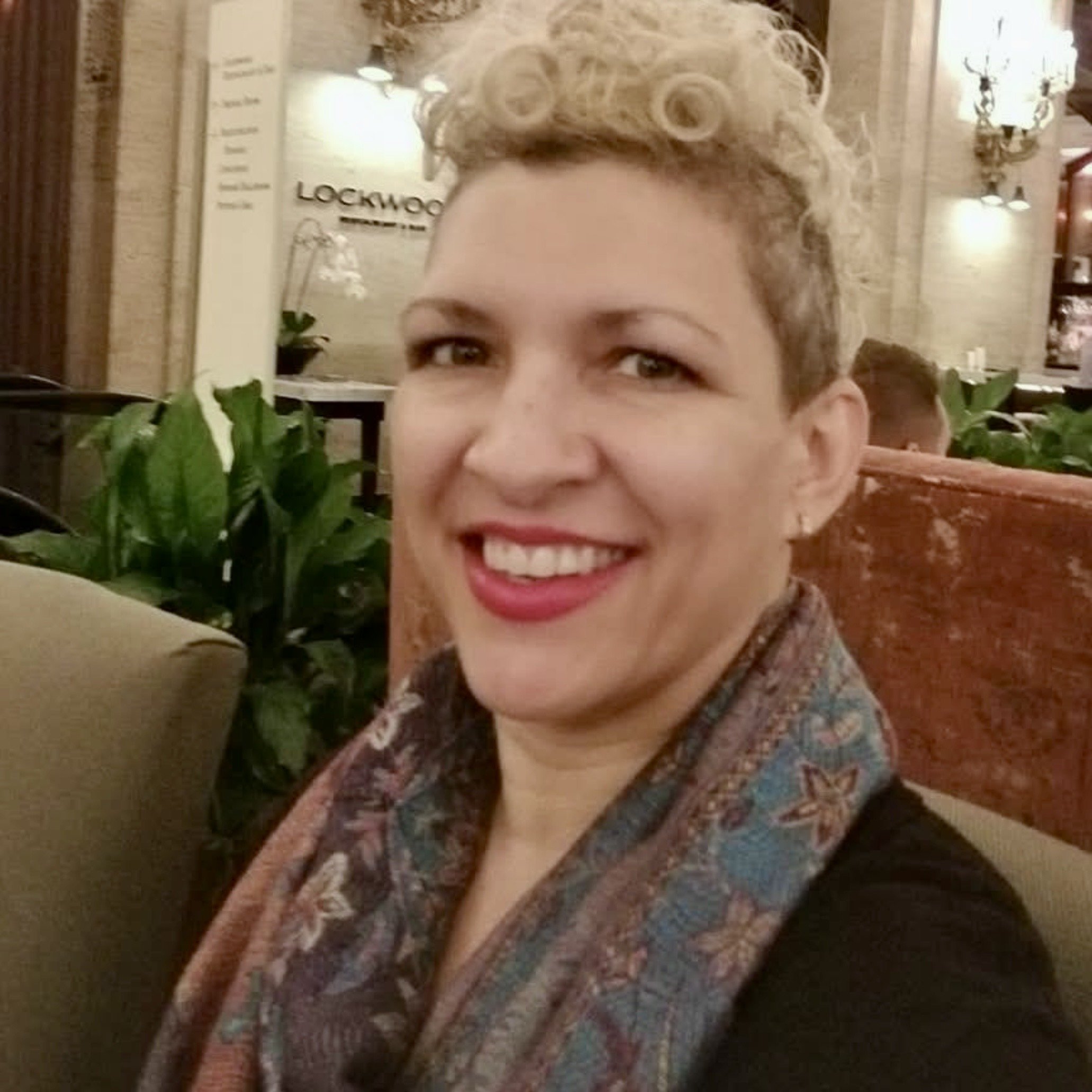 A woman with curly white hair styled in a high updo, wearing red glasses, a red shirt, and a multicolored scarf, sitting inside a vehicle and smiling.