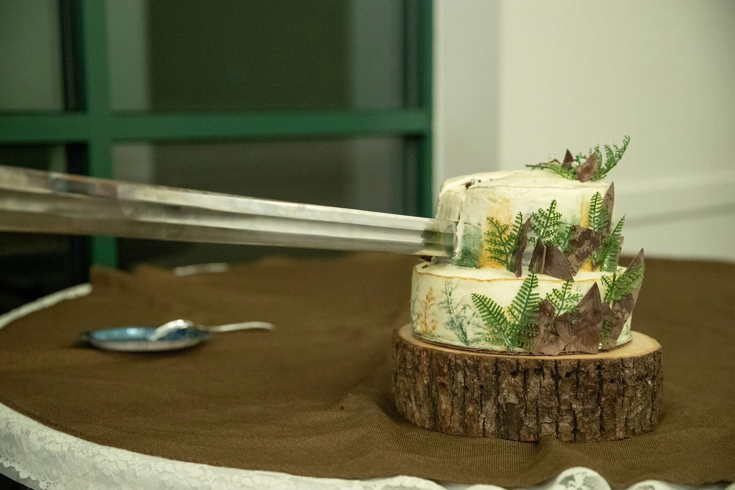 A two-tiered cake decorated with green fern leaves and dark brown edible decorations sits on a slice of tree trunk on a table. Tongs are cutting into the top of the cake.
