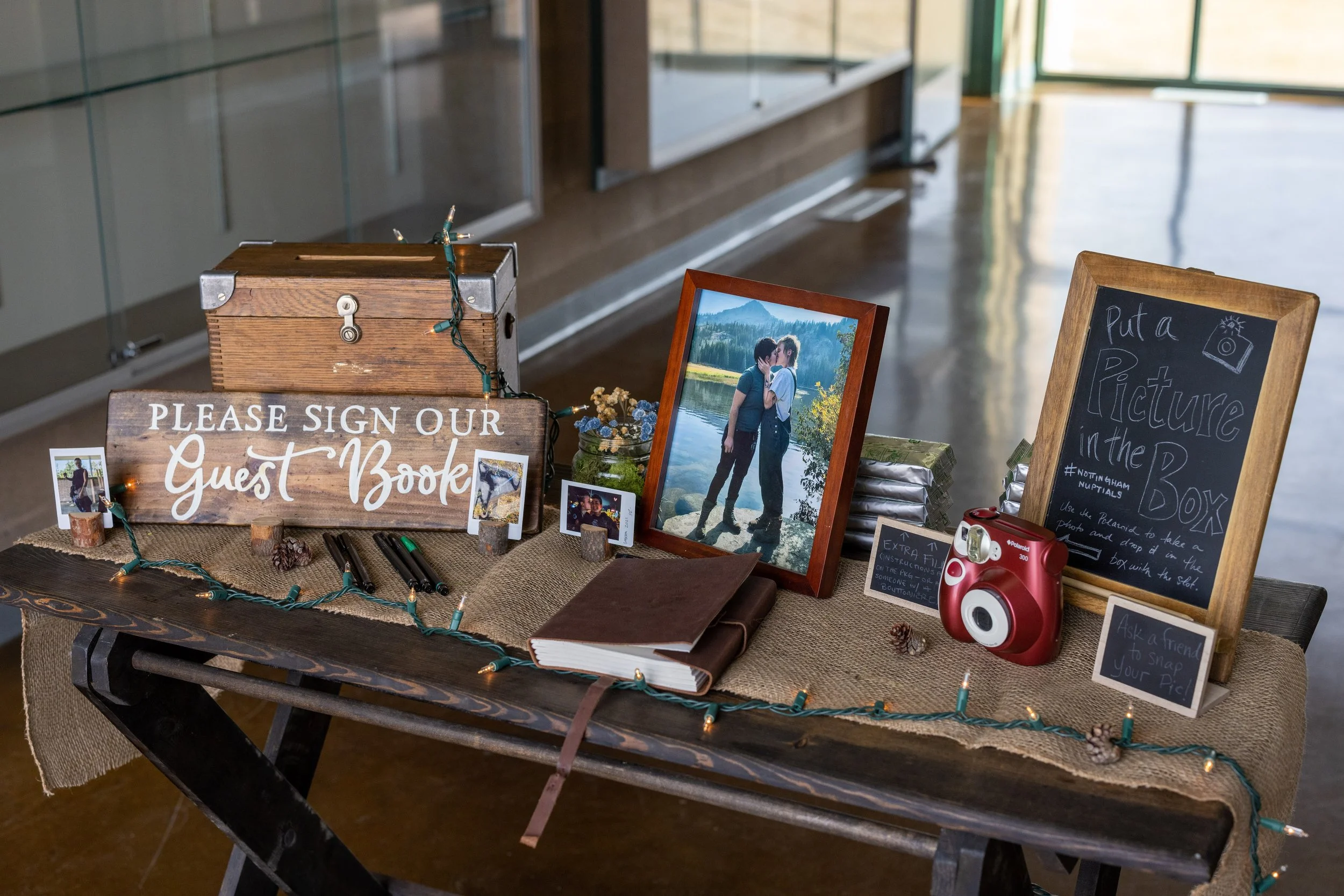 A table with a sign asking guests to sign the guest book, a framed photo of two people kissing by a lake, a chalkboard sign for a picture box, a red instant camera, and small framed photos, all decorated with string lights and pinecones, set up for a