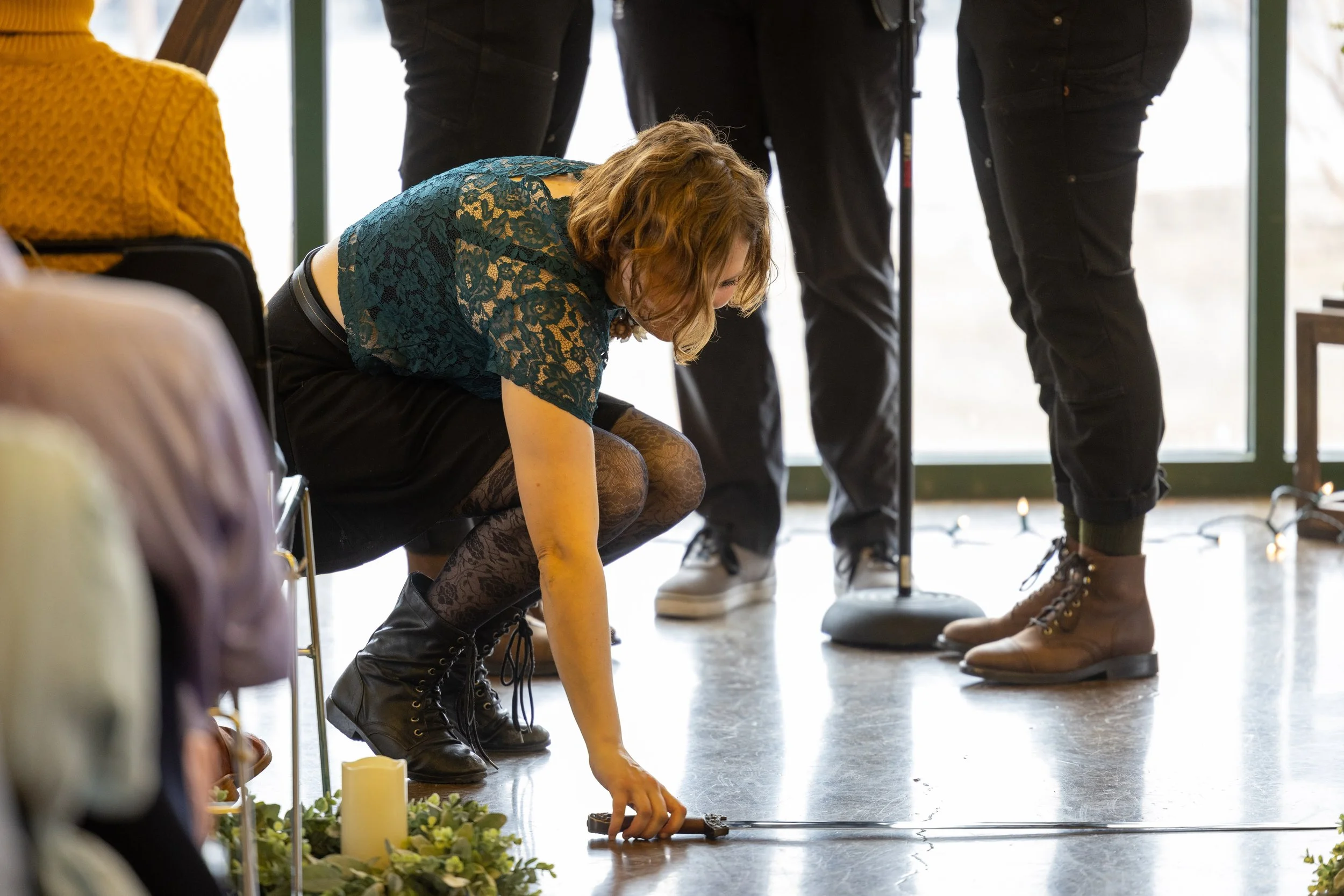 Woman placing a candle on the floor during a ceremony, surrounded by seated and standing people indoors near a large window.