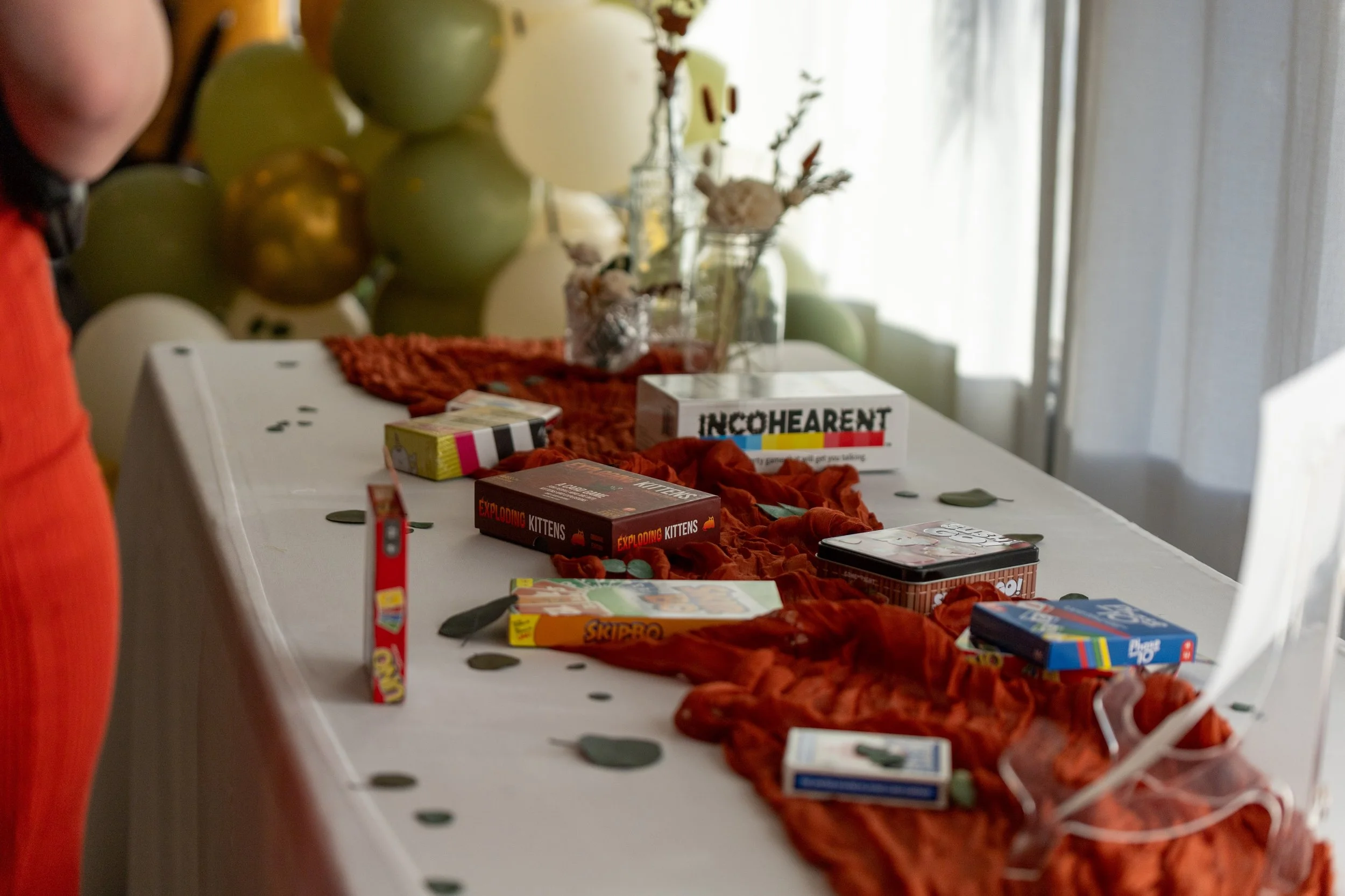A decorated table with board games, snacks, a red table runner, and balloons in the background for a celebration.