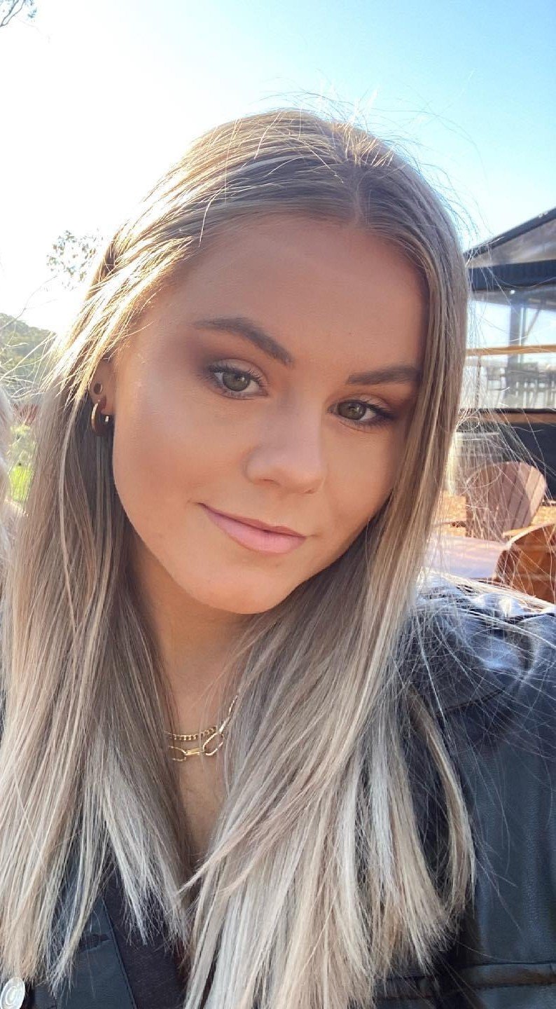 A woman with blonde hair styled in a messy bun and loose curls, smiling, wearing a beige blazer and a matching top, standing against a plain white background.