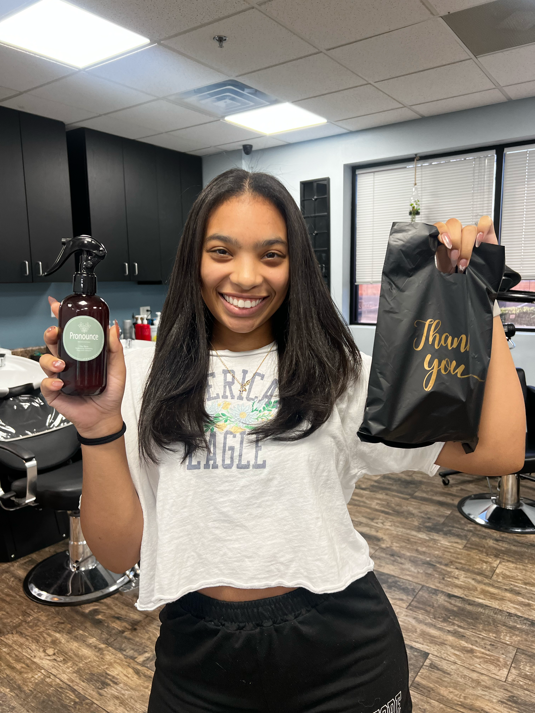 Happy woman holding a spray bottle labeled 'Pronounce' in her left hand and a black gift bag with 'Thank you' written on it in her right hand, inside a salon or similar setting.