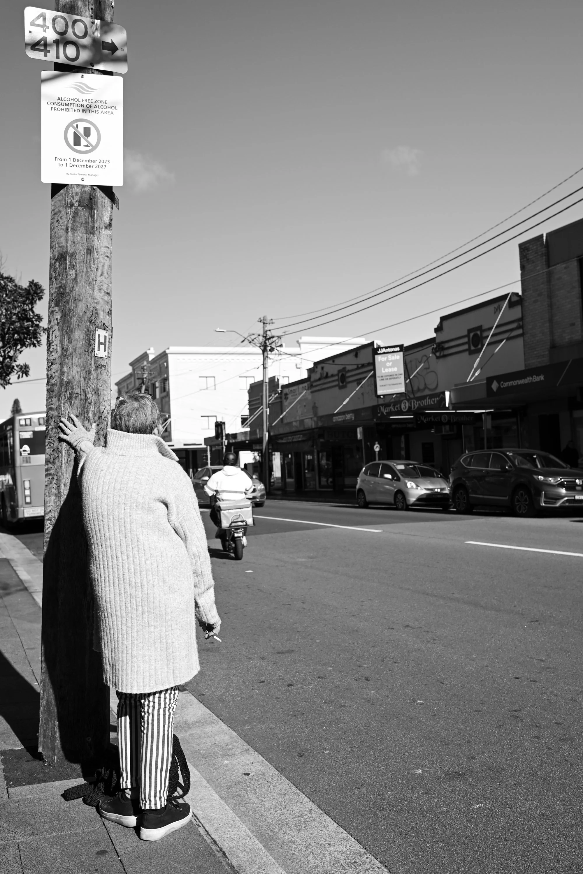 Black-and-white street photograph in Randwick, Sydney, showing a person from behind leaning one hand against a wooden utility pole beneath bus stop and alcohol-free zone signs, while traffic and shopfronts stretch along the road under a clear winter 