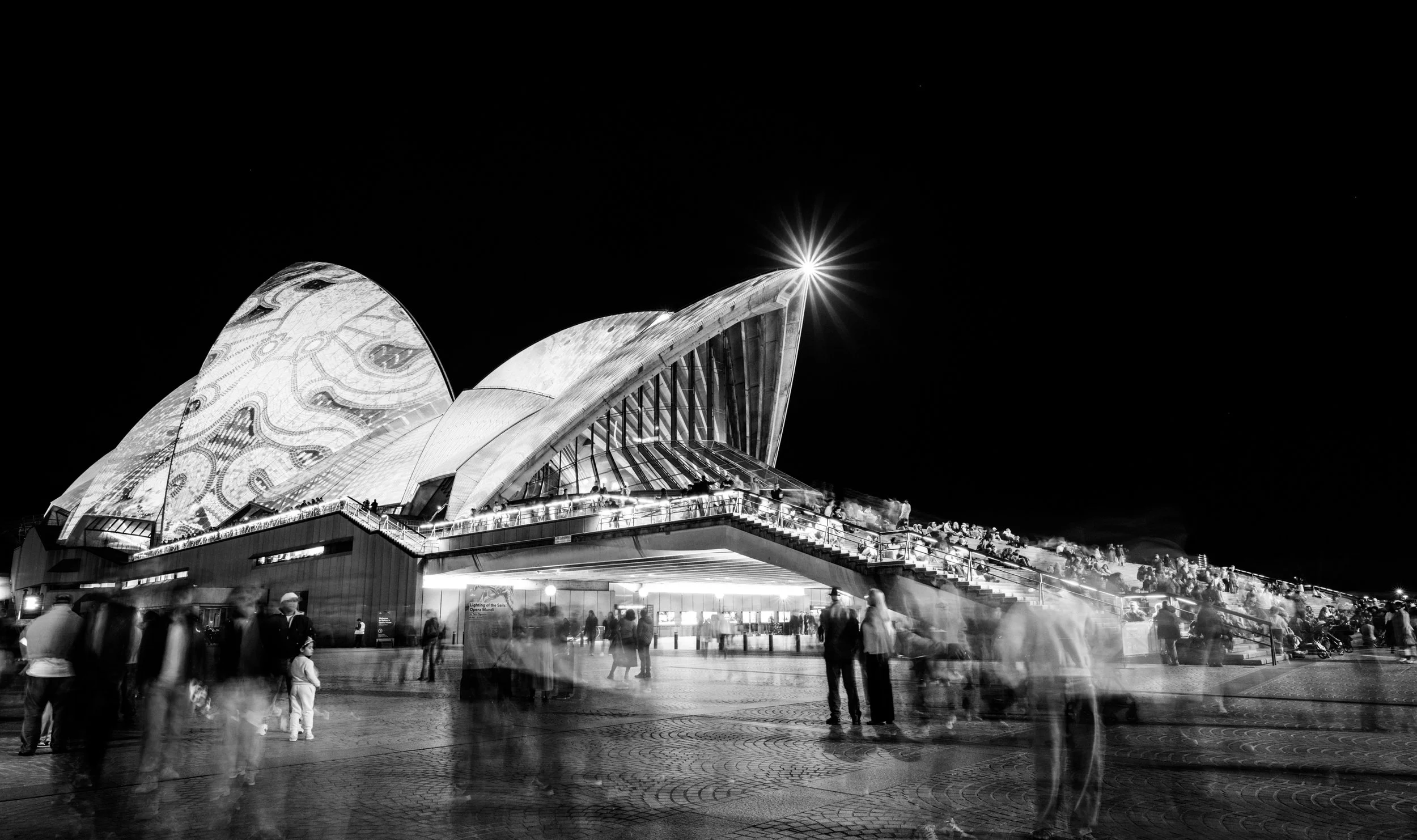 Black and white long-exposure photograph of the Sydney Opera House during Vivid Sydney, June 2026. Crowds move across the forecourt beneath illuminated sails, creating a dense field of blurred bodies, light, walking, and shared urban presence.