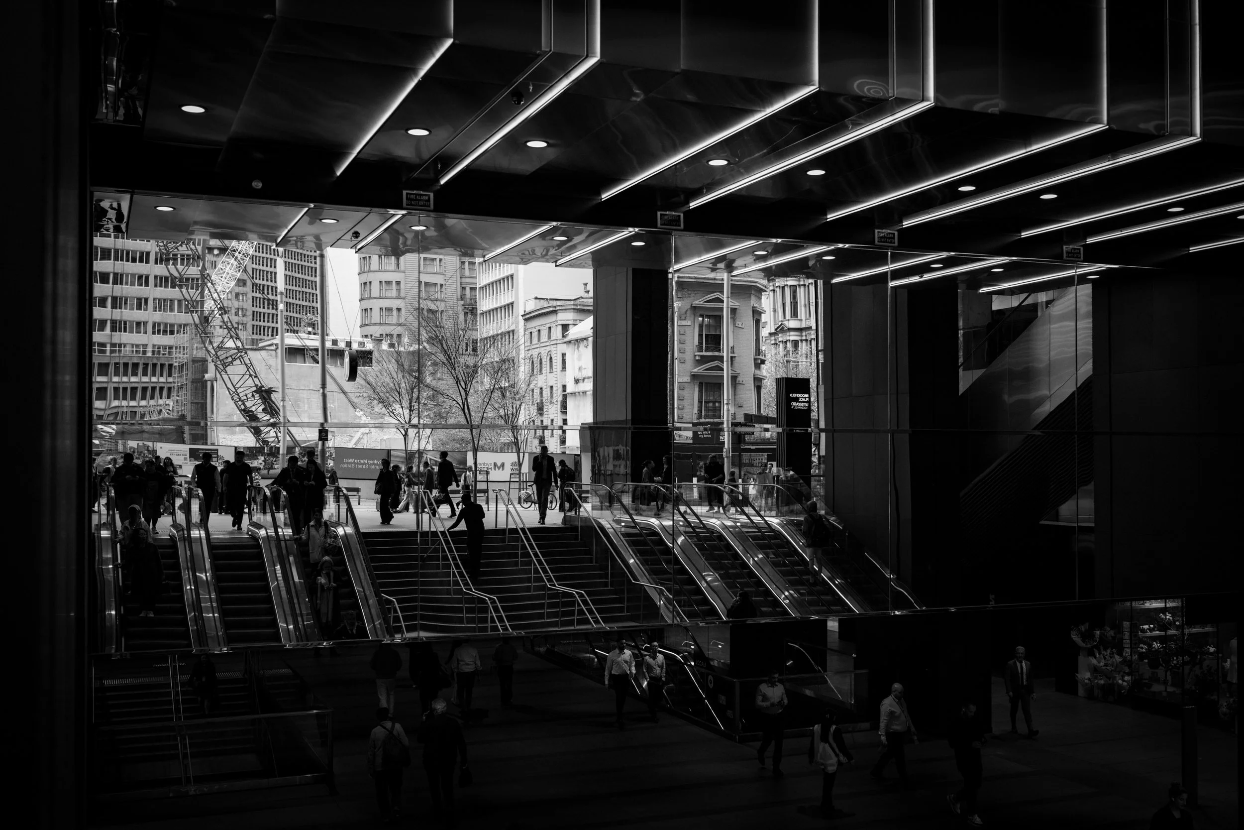 Black and white architectural photograph of the George Street entrance to Wynyard Station at Brookfield Place Sydney / Wynyard Place in Sydney CBD. Commuters move through escalators, stairs, glass, shadow, reflected surfaces, and a small T station si