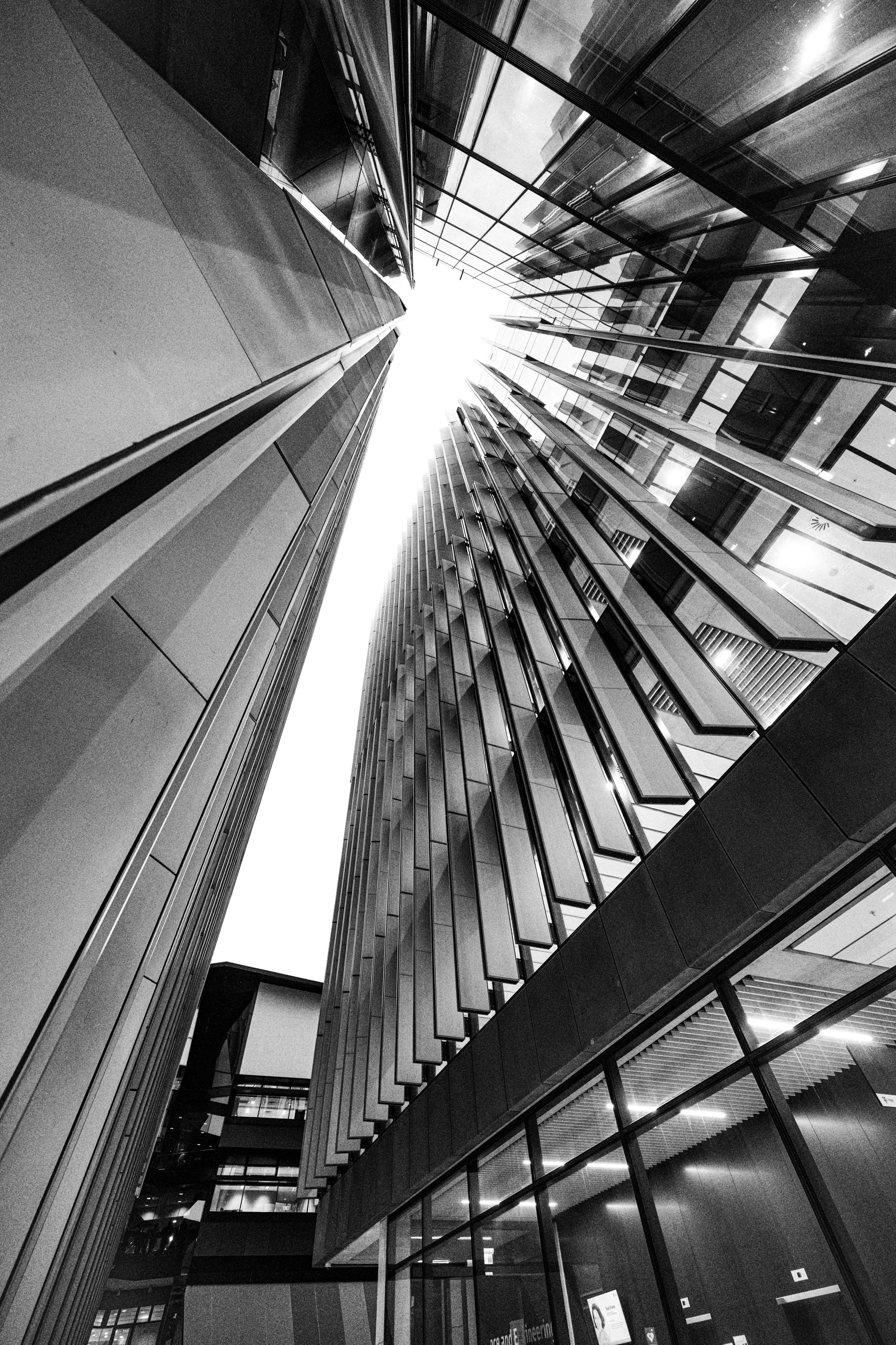 Black-and-white architectural photograph looking sharply upward through a narrow passage at UNSW Kensington Campus in Sydney, Australia. Tall glass and concrete building façades rise on both sides, forming a dramatic vertical corridor or portal of li