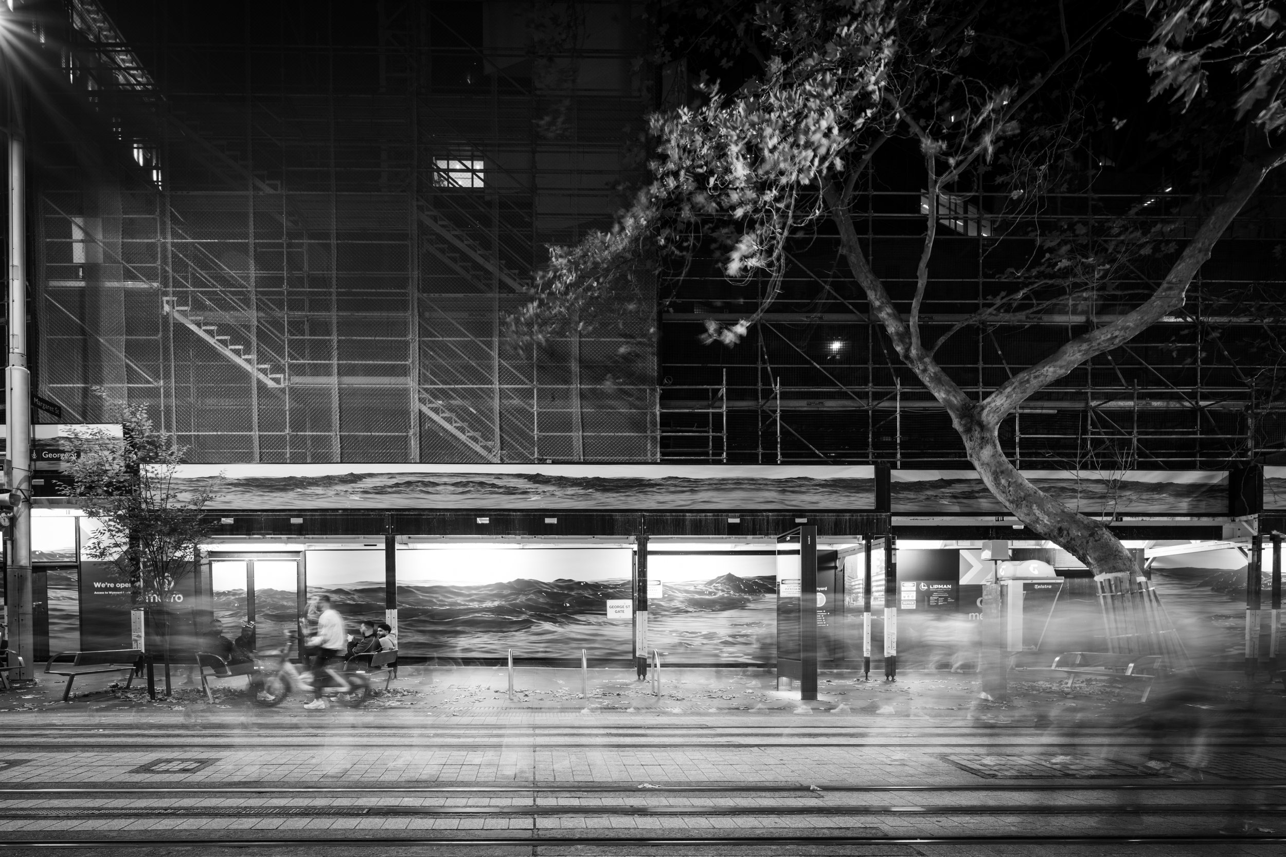 Black-and-white night photograph of George Street, Sydney, with light rail tracks, blurred pedestrians, scaffolding, a tree, and Ash Garwood’s sea artwork Equivalence on a construction hoarding.