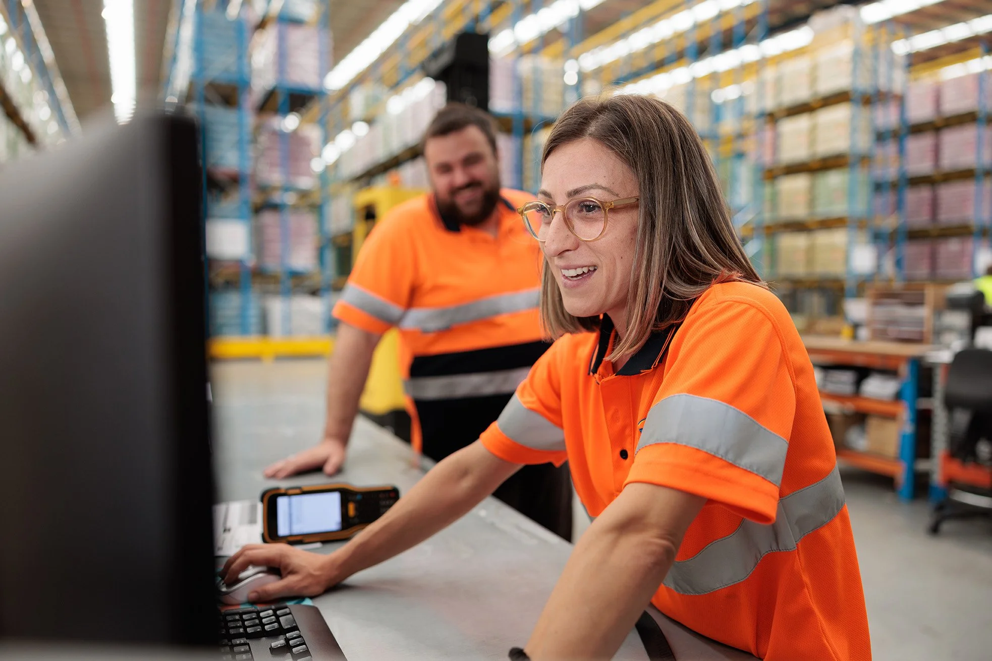 Female warehouse worker with glasses in orange safety uniform working on a computer in a warehouse, with male worker in the background smiling.