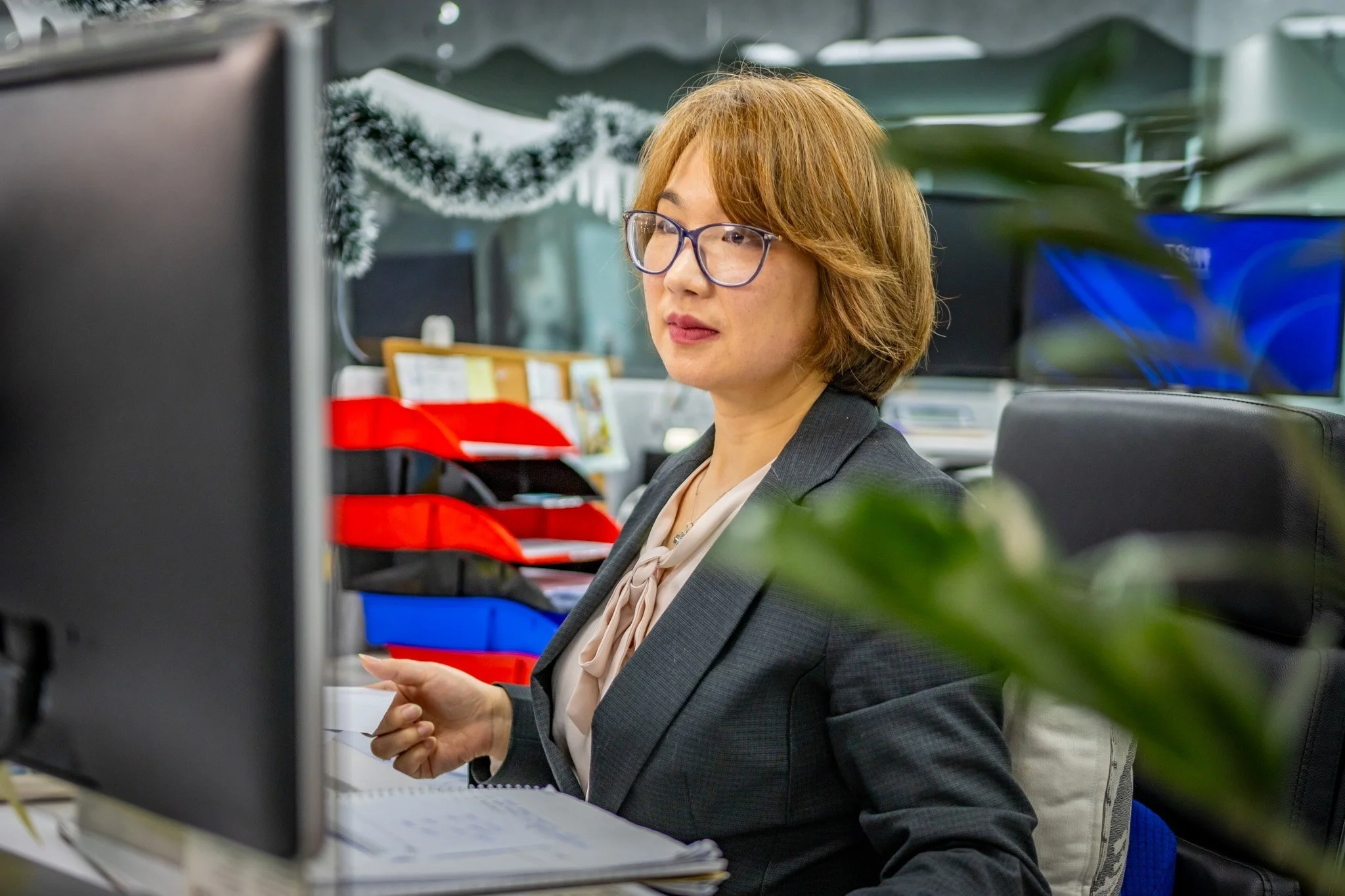 A woman with glasses, reddish hair, and a blazer is working at a desk with office supplies, surrounded by monitors and plants.