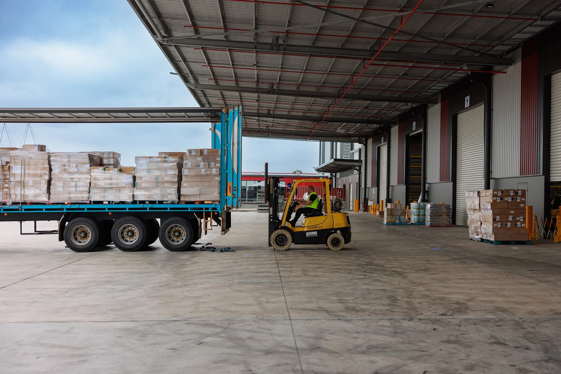 A warehouse loading area with a forklift operator moving pallets of boxed goods near a truck with more pallets on it. The warehouse has a large covered space with multiple storage doors and various equipment.