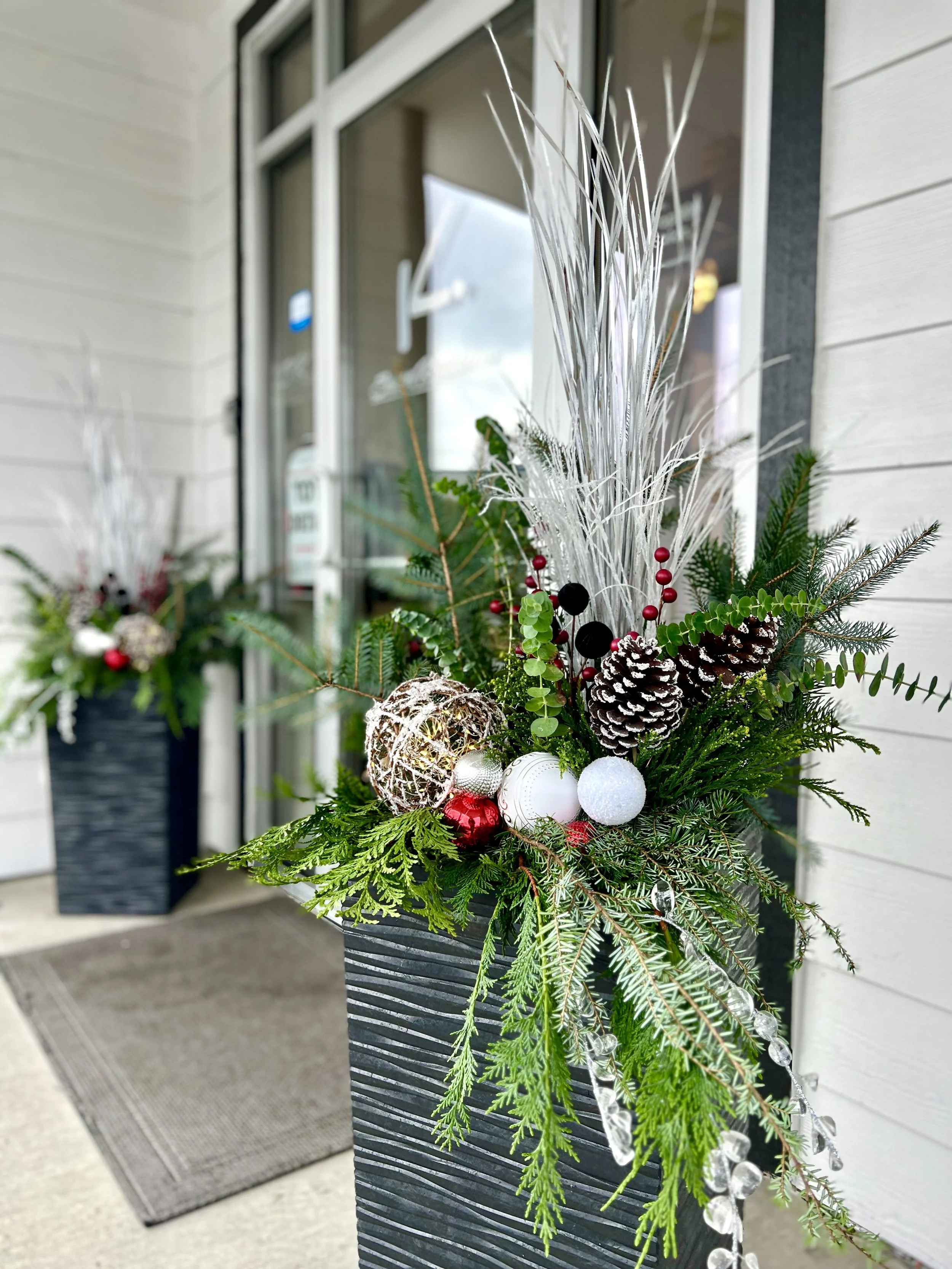 Decorative holiday floral arrangement with pinecones, ornaments, and greenery in a black planter on a porch.