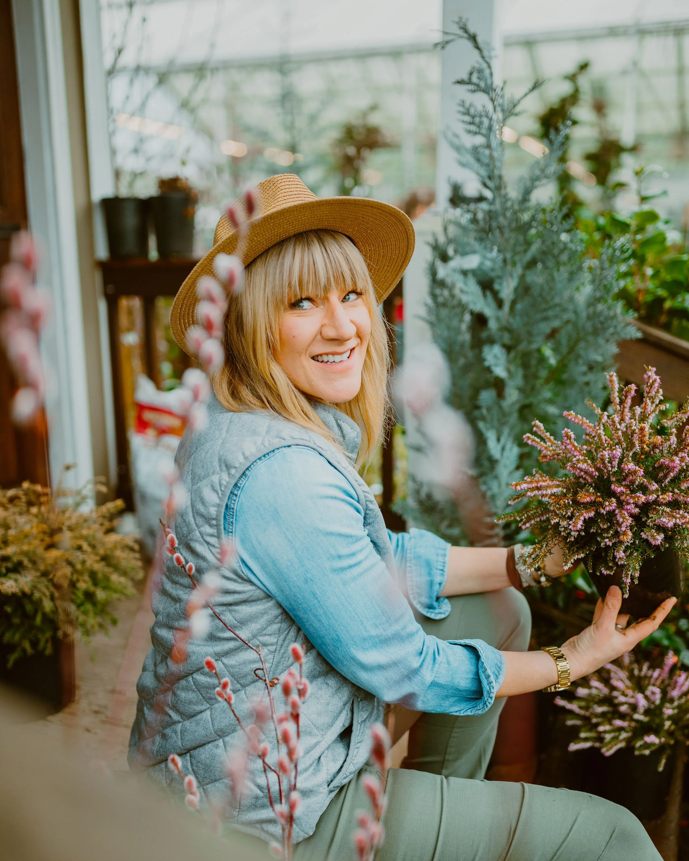 A woman with blonde hair wearing a straw hat, denim vest, and khaki pants, smiling at the camera while holding a potted purple heather plant inside a greenhouse surrounded by other plants and flowers.