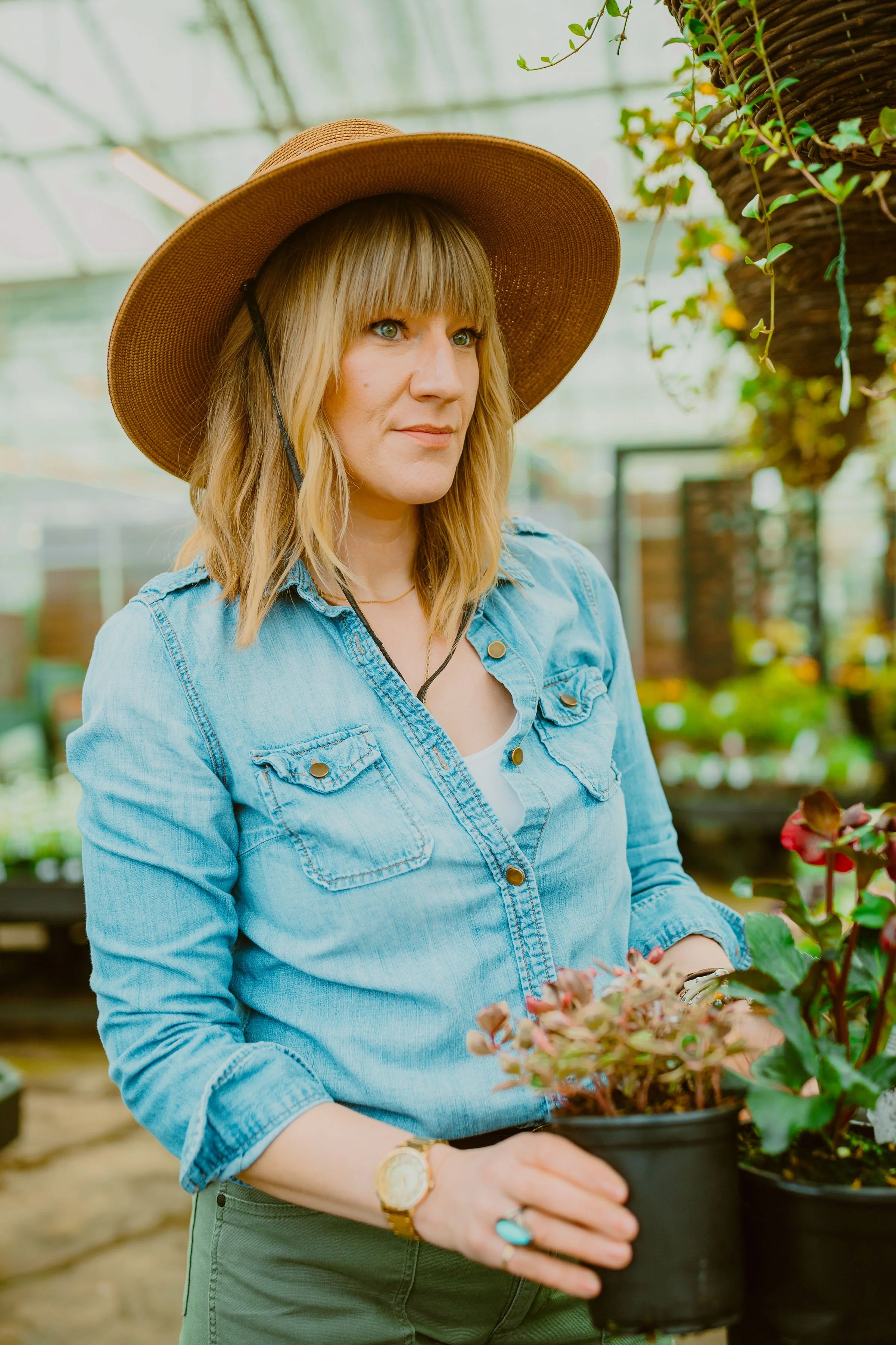 A woman with blonde hair wearing a wide-brimmed straw hat and a light blue denim shirt, holding a potted plant in a greenhouse filled with various plants and greenery.