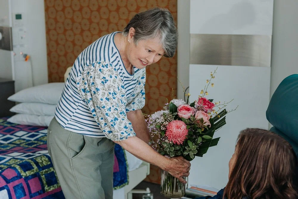 An elderly woman gives a bouquet of pink and white flowers to a woman sitting in a hospital bed.