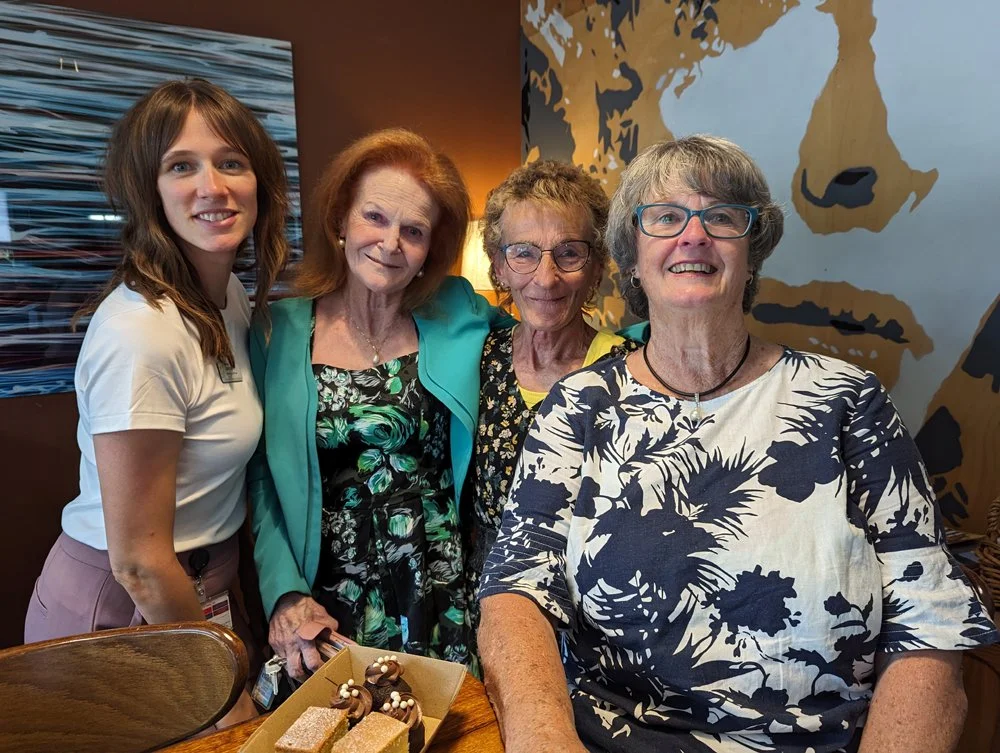 Four women gathered around a table, smiling; one woman is in the foreground and three women are in the background, standing against a decorated wall.