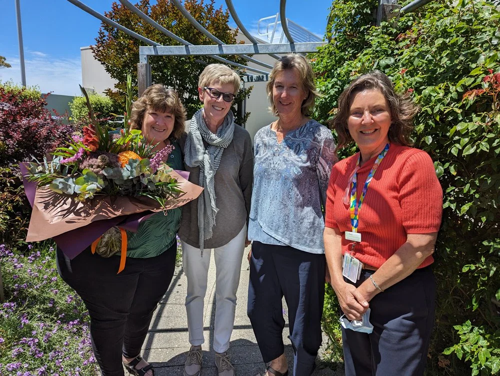 Four women standing outside in a garden, smiling at the camera. One woman is holding a large bouquet of flowers. Blooming plants and a clear blue sky are visible in the background.