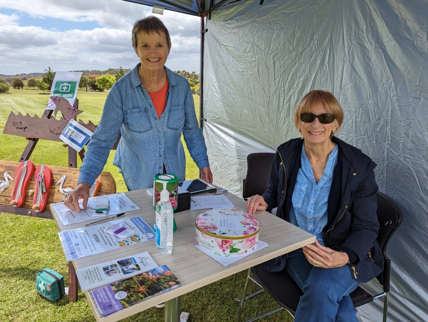 Two women at a stall outdoors under a canopy, with tables, flyers, and medical supplies. One woman is standing, smiling, wearing a blue denim shirt; the other is seated, smiling, wearing sunglasses and a dark jacket. There are decorative wooden swans