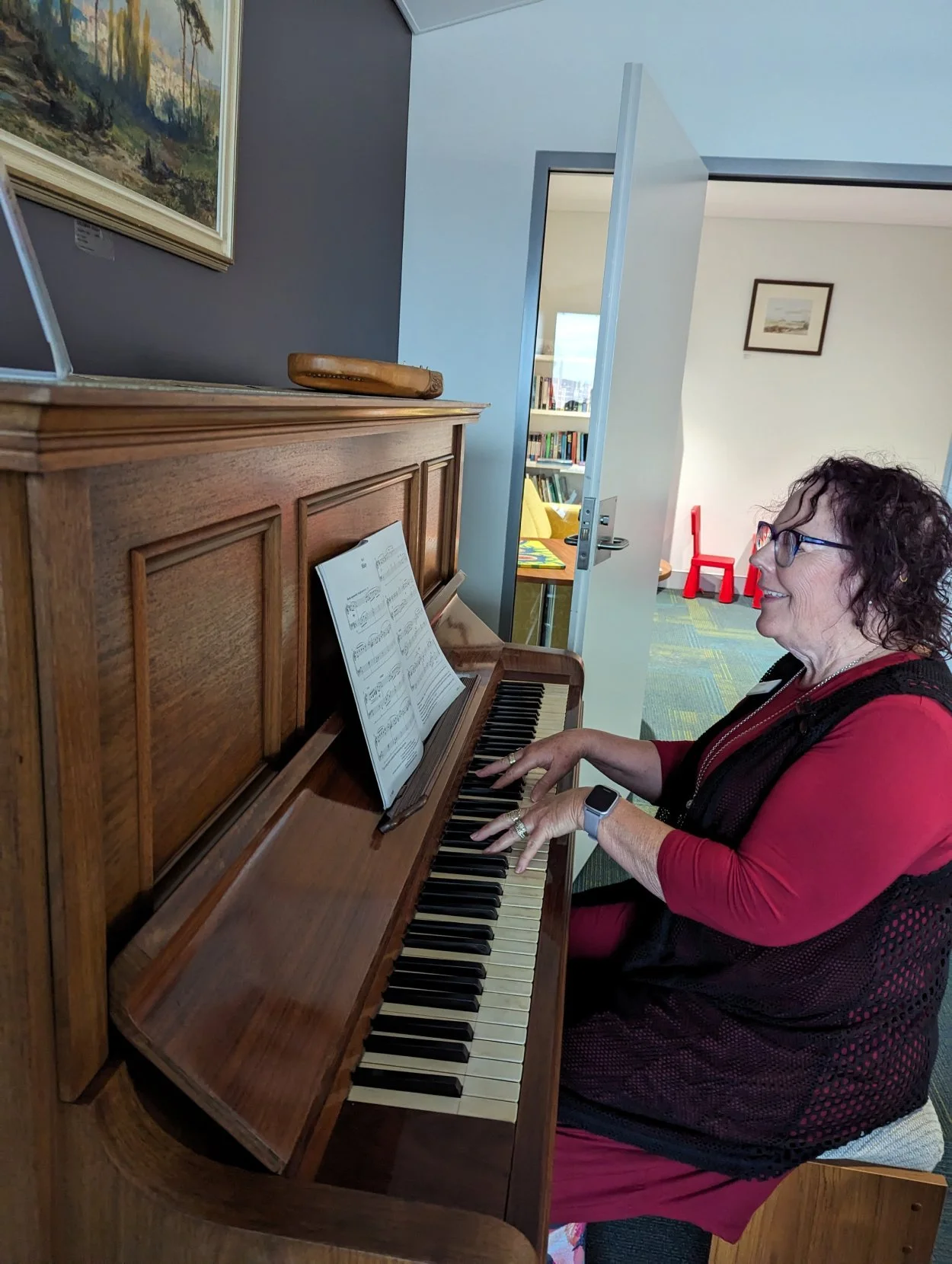 A woman with curly hair and glasses playing a wooden upright piano with sheet music on the stand.