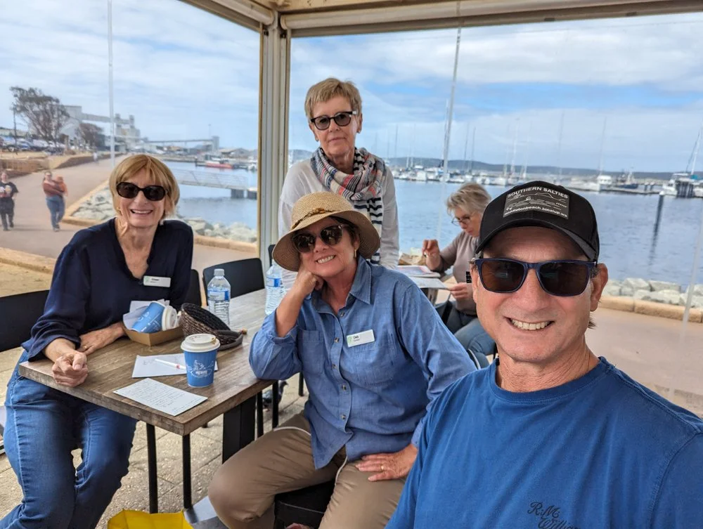 A group of five people enjoying a day at the waterfront restaurant, with docked boats and a cloudy sky in the background. They are sitting and standing around a table with drinks and a menu, smiling for the photo.