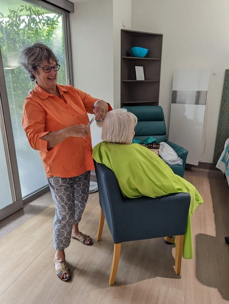 A woman with gray hair, glasses, and an orange shirt is cutting another woman's hair in a salon or home setting. The woman receiving the haircut has white hair and is seated in a blue chair, covered with a bright green cape. There is a large window w