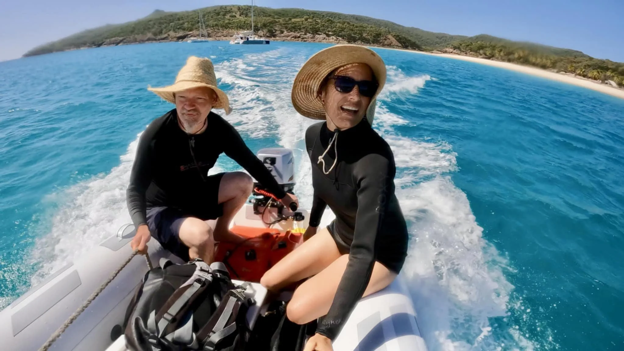 Two people on a boat in turquoise water, wearing wide-brimmed hats and sunglasses, enjoying a sunny day near a coastline with green hills and a sandy beach.