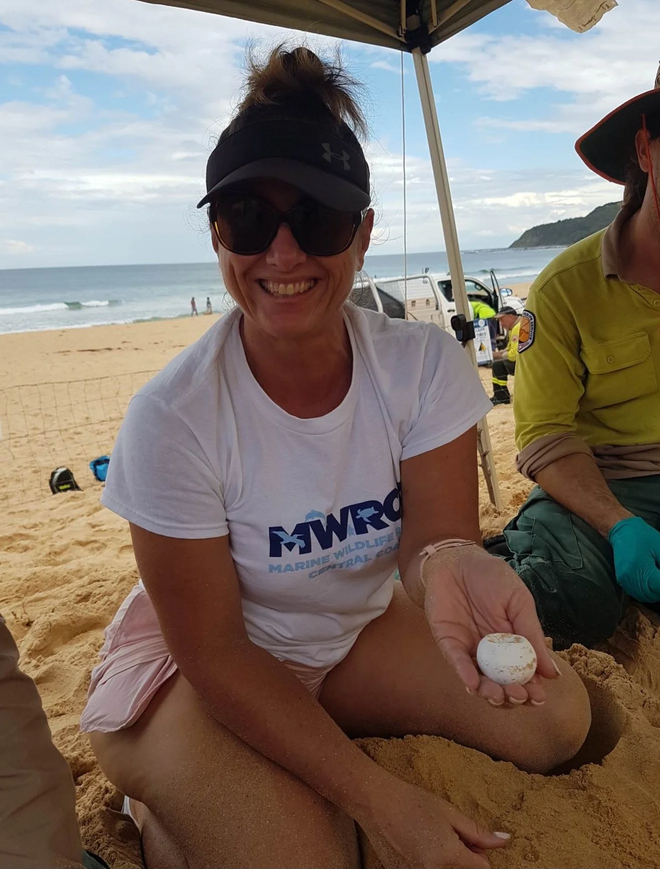A woman wearing sunglasses, a black visor, and a white t-shirt with the MWRC logo, sitting on the sand at the beach, holding a white egg or shell, smiling at the camera.