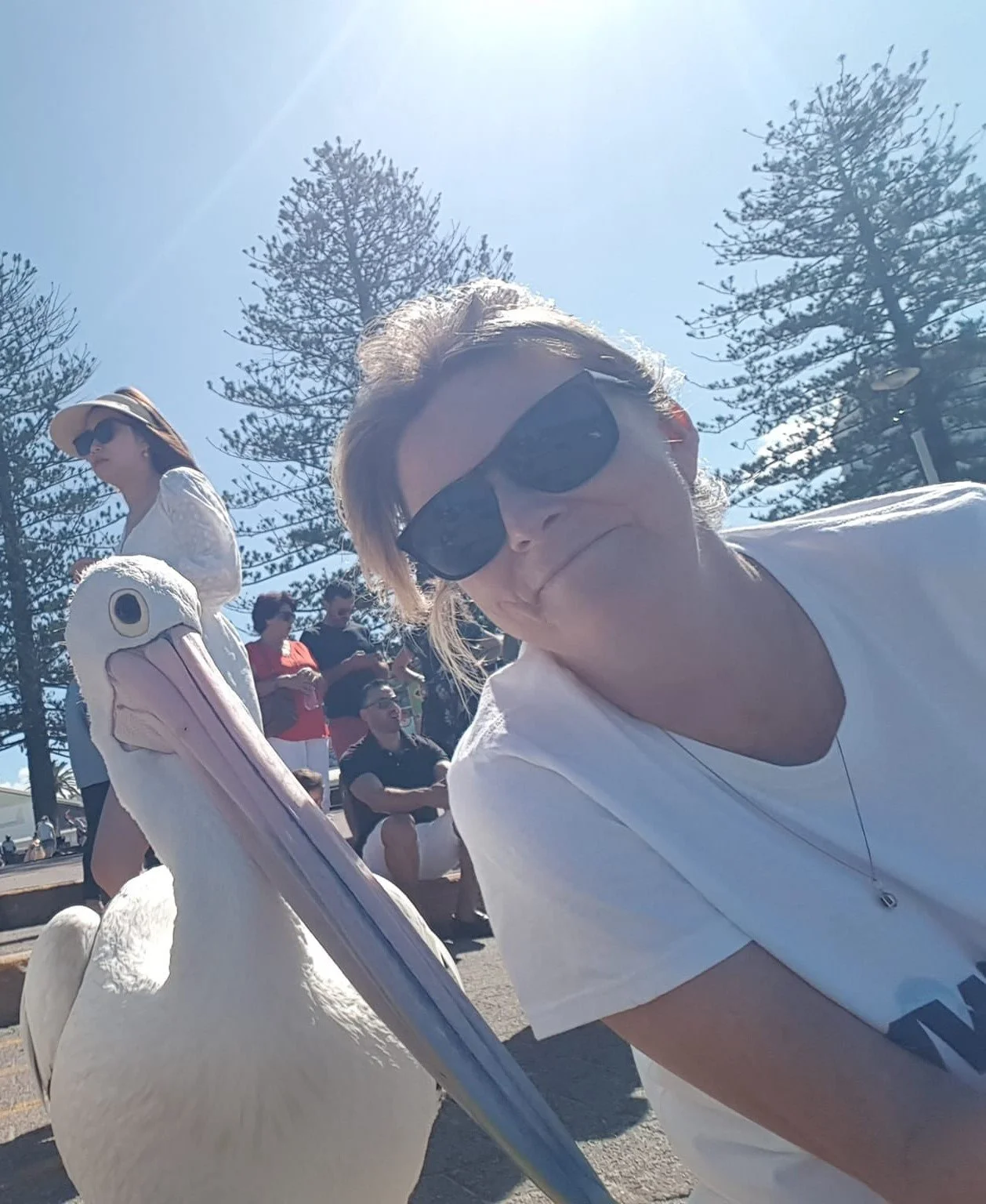 A woman wearing sunglasses and a white T-shirt taking a selfie with a pelican on her shoulder during a sunny outdoor event, with other people and trees in the background.