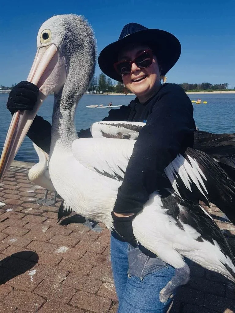 Woman in sunglasses and a wide-brimmed hat hugging a large pelican outdoors near a body of water on a sunny day.