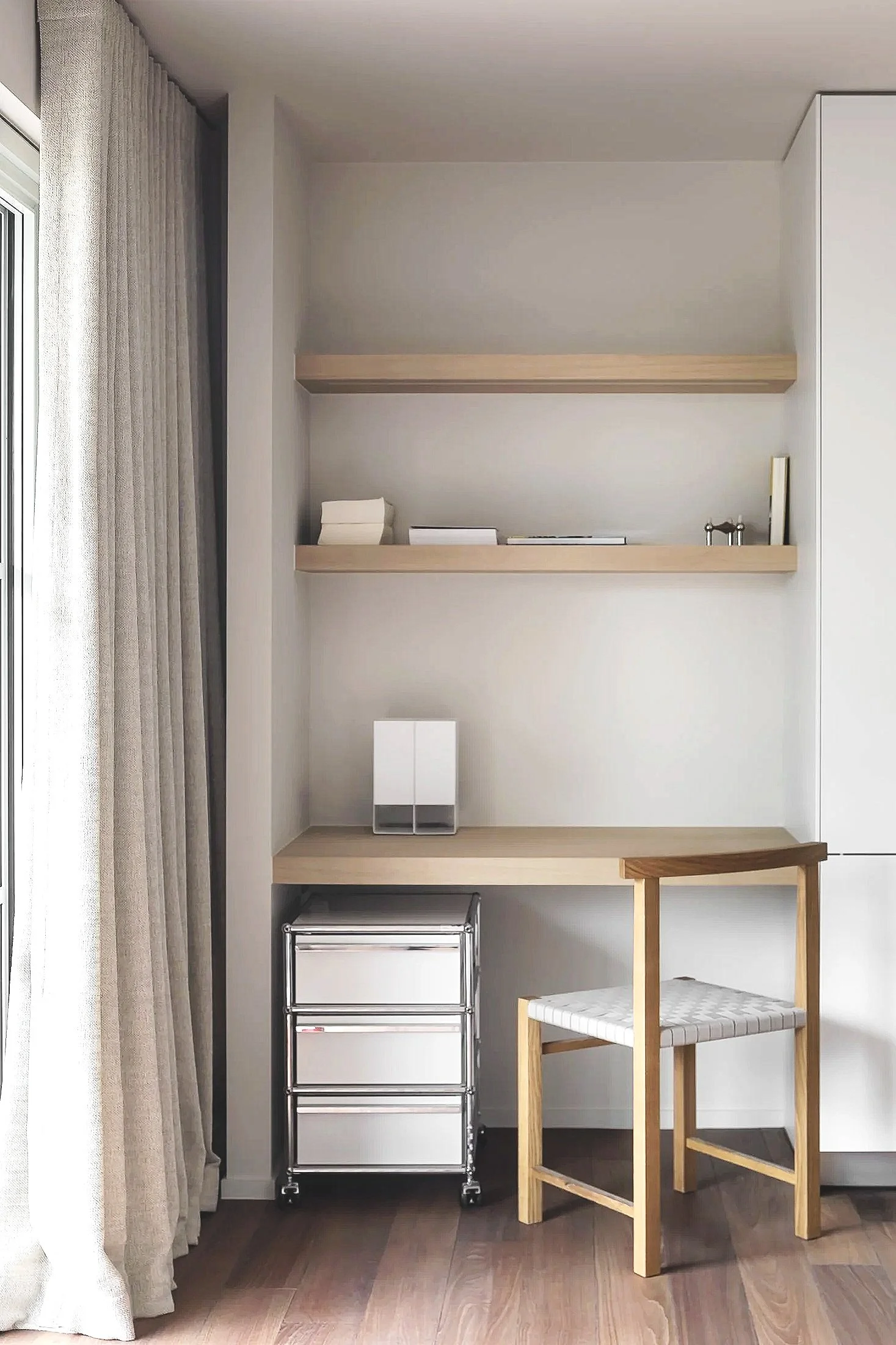 A minimalist corner with beige curtains on the left, two wooden floating shelves on the wall holding a few objects, a small metal storage cart with four drawers, a tall white cabinet on the right, a low table or desk with a wooden frame and a white cushioned seat, and a wooden chair with a cushioned seat in front. The flooring is light-colored wood.