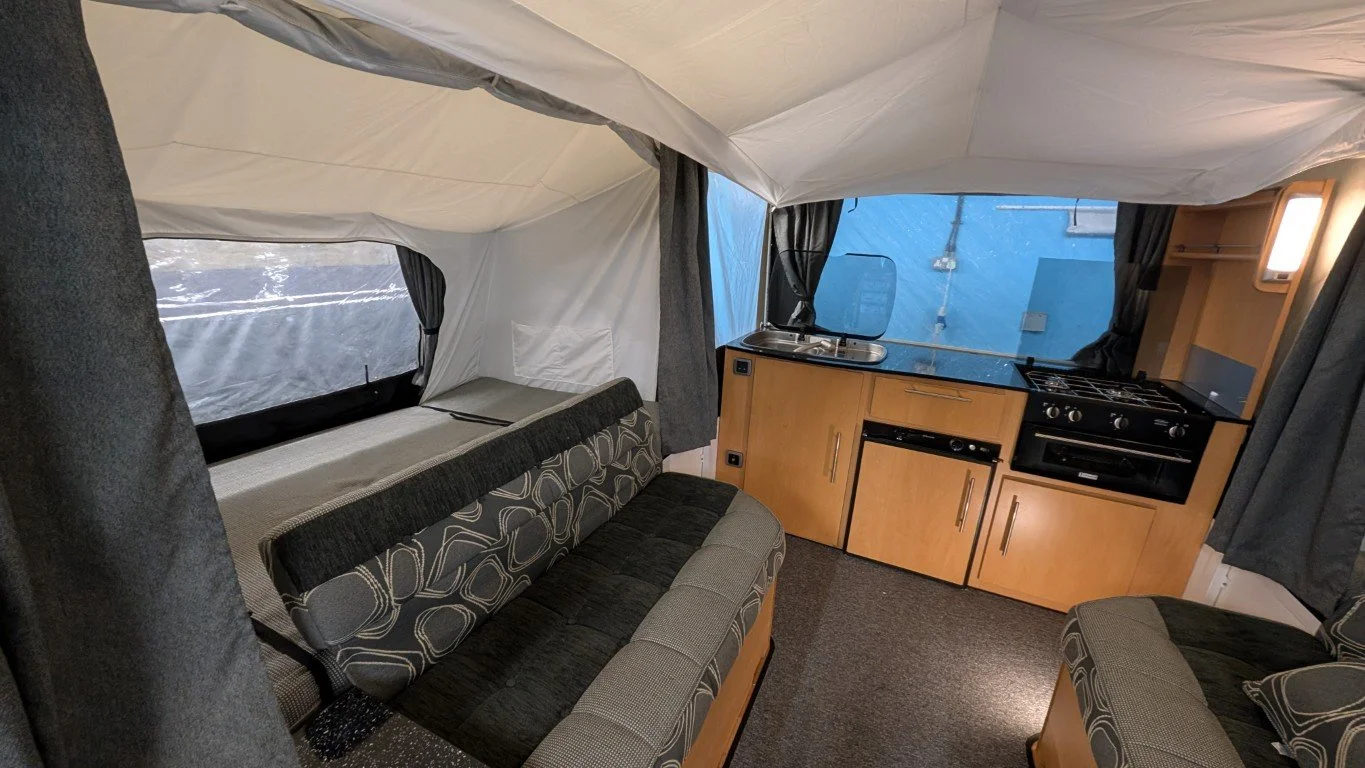 Interior of a small camper with a cushioned seating area, a window with a curtain, and a kitchenette with a sink, stove, and cabinets.