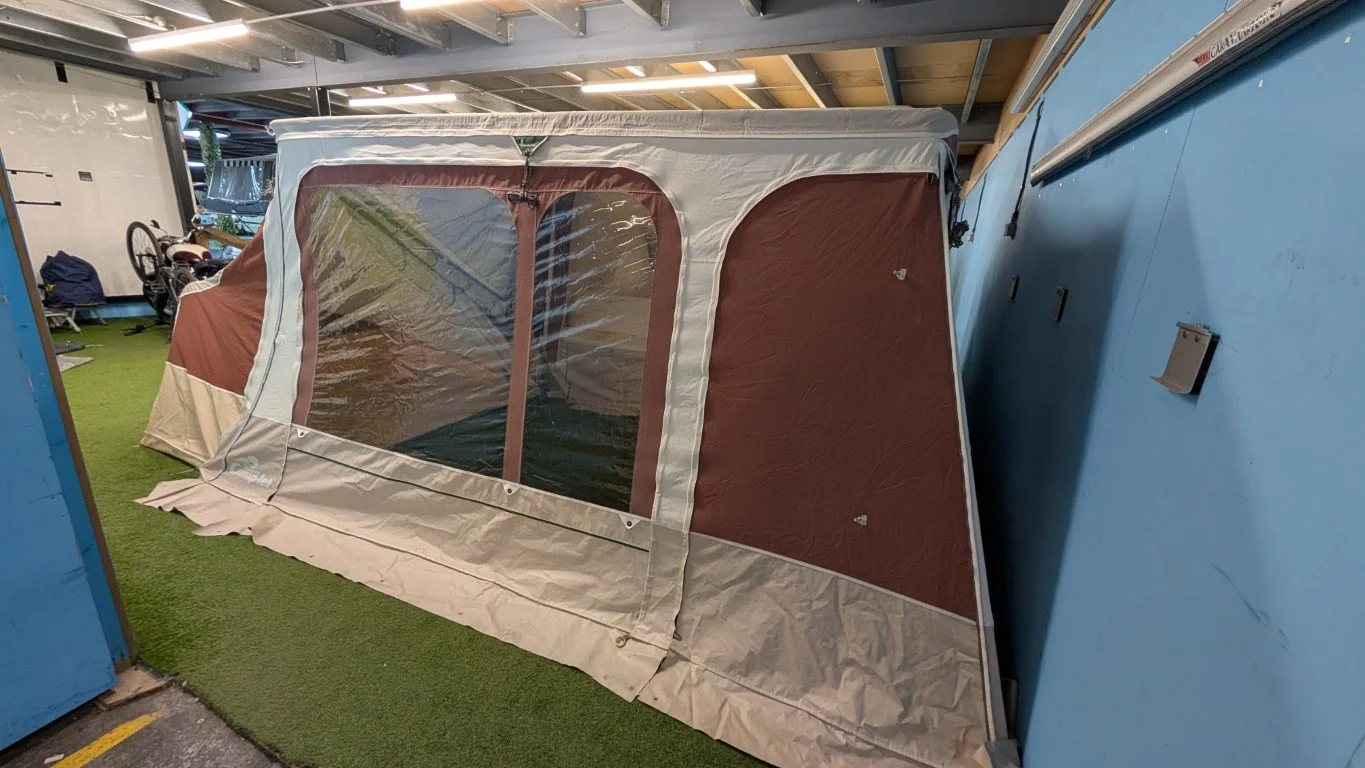 A portable camping tent with brown and beige fabric, large clear plastic windows, set up indoors on artificial grass flooring, next to a blue wall with hooks.