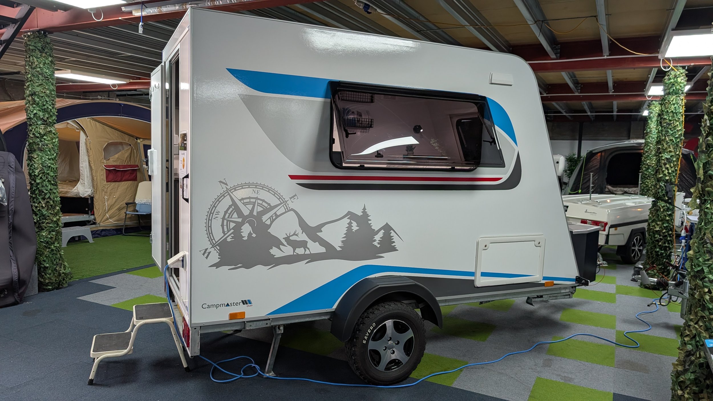 A white travel trailer with mountain and forest landscape graphic on the side, inside an indoor showroom with tents, greenery, and other RVs visible. The trailer has a window and appears to be part of an exhibit or sales display.