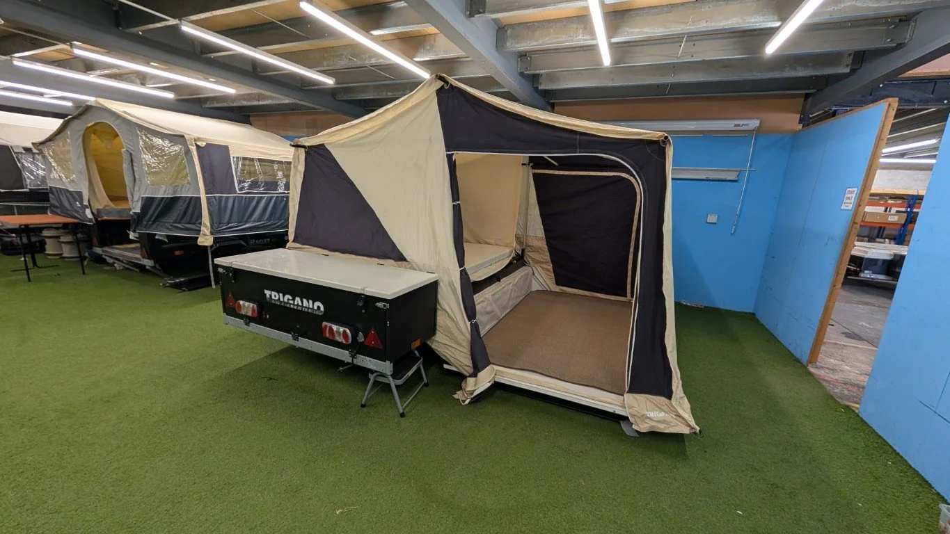 A beige and black tent set up inside a retail store, with its sides open and a mattress inside, attached to a small black trailer labeled 'TIGER'. The store has artificial lighting and a green carpeted floor.