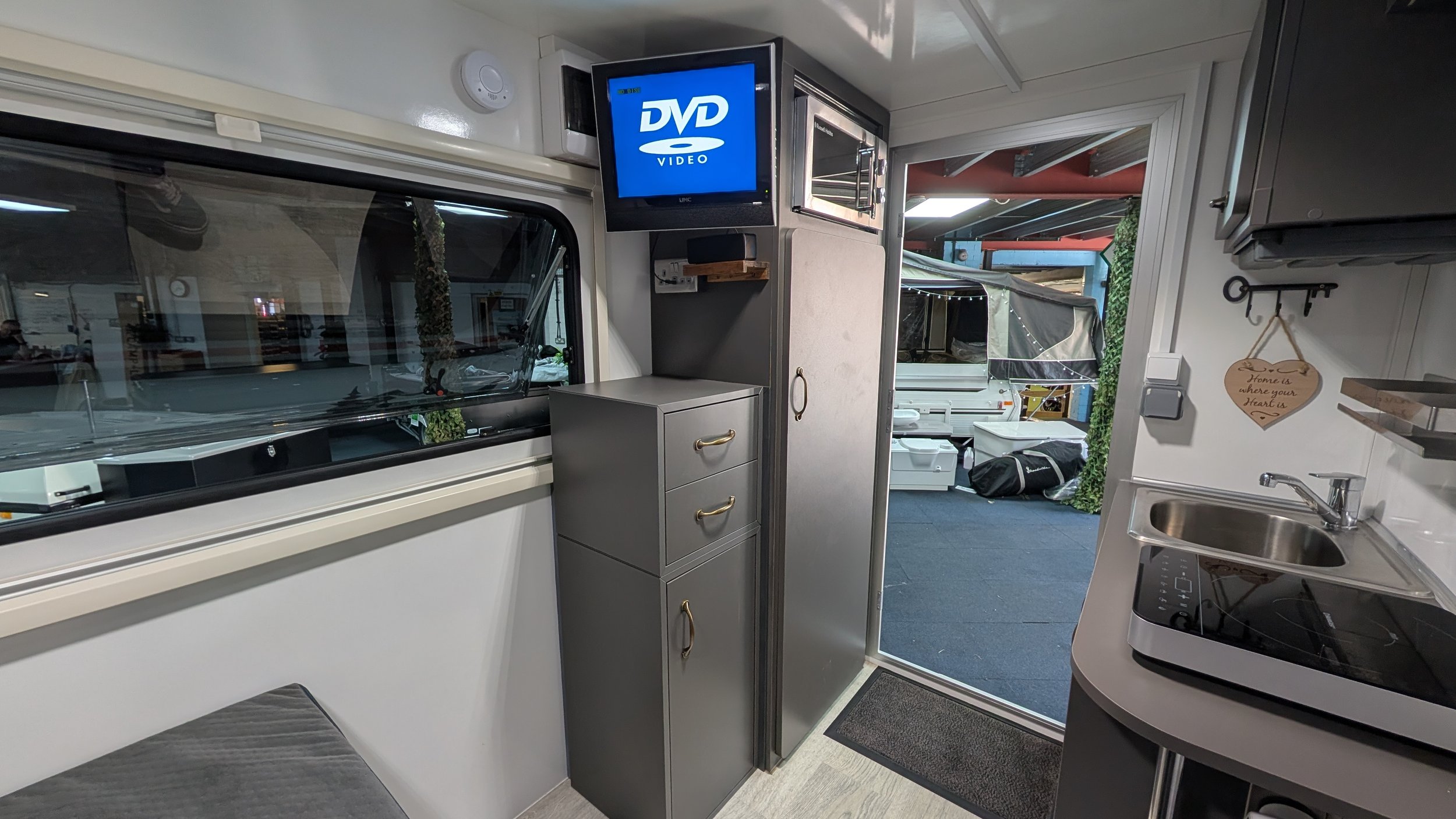 Inside a camper van, showing a small kitchen area with a sink, a portable stove, and cabinets. A small window is visible on the left, and outside the door, there is a covered outdoor space with some outdoor equipment and a black bag.