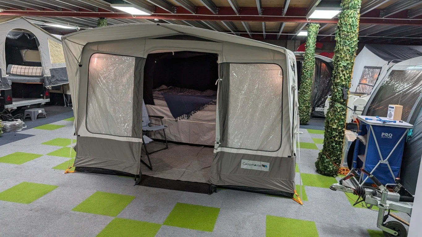 Portable camping tent set up indoors, with a bed and chair inside, surrounded by other tents and camping equipment on a checkered green and gray carpet.