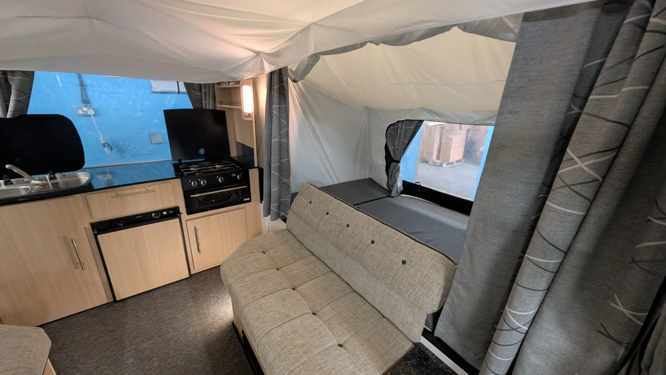 Interior of a compact camper with a small kitchenette, a beige sofa, and a sleeping area with windows.