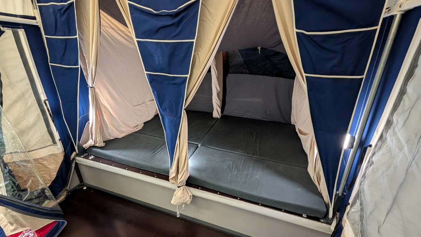A camping tent with open blue and beige fabric flaps, revealing a black mattress inside, set up on a dark floor.