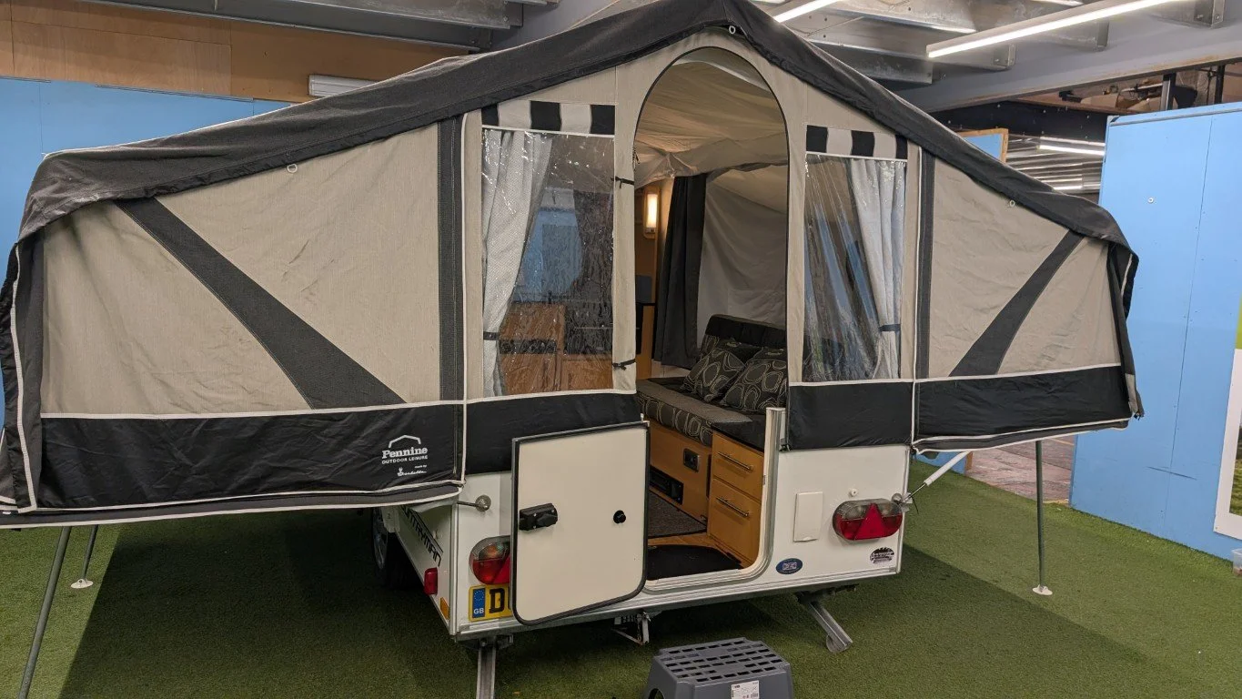 Pop-up camper trailer with extended sleeping tents on both sides, open door revealing interior with gray and black patterned cushions and cabinets, set on artificial grass in a showcase area.