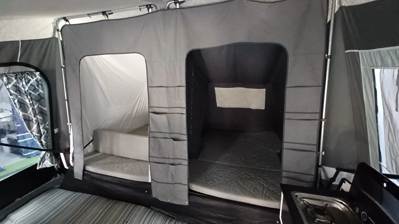 Inside a Camp-let Trailer Tent with a sleeping area that has a set of bunk beds, a small bed or mattress, and windows with curtains.
