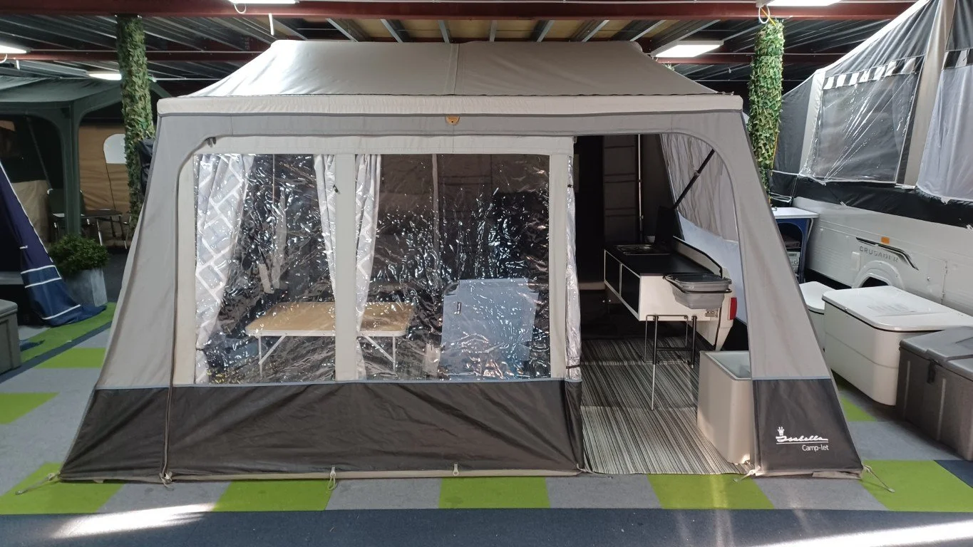 A Camp-let Trailer Tent set up indoors, with clear plastic windows, a small kitchen unit with a stove and sink, and a striped rug on the floor. There are camping accessories and other tents in the background.